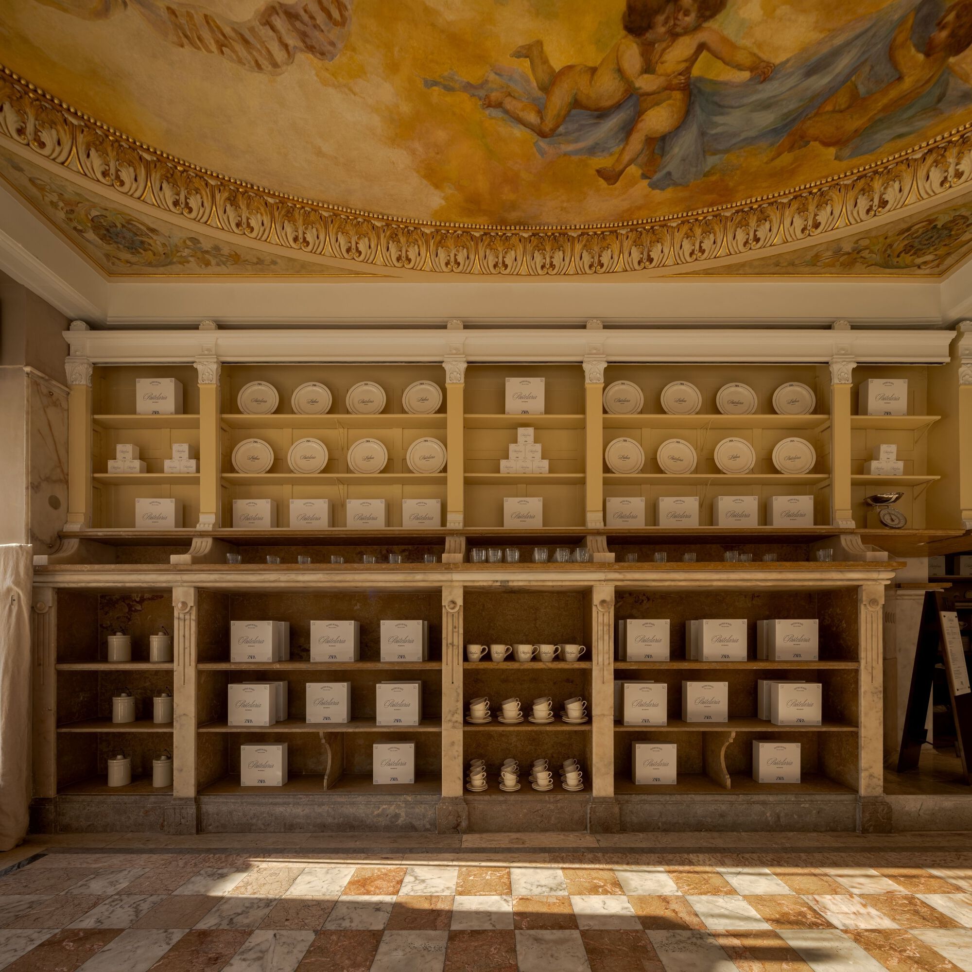 Built-in display shelving with white boxes and plates beneath a painted dome ceiling with gilded molding