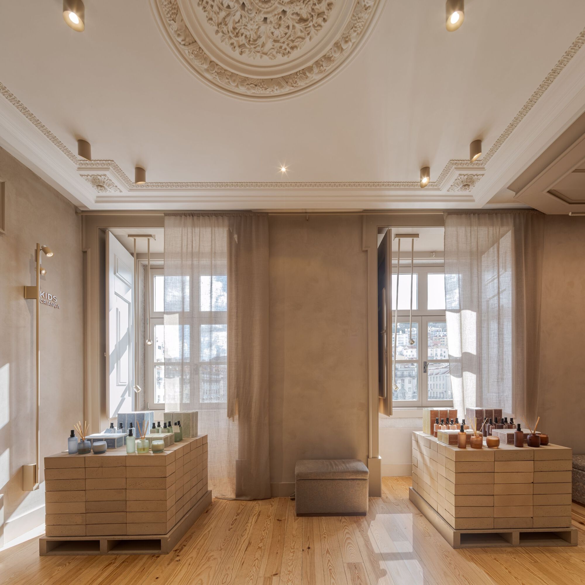 Symmetrical room with timber product displays beneath tall windows dressed with sheer curtains and ornamental ceiling rosette