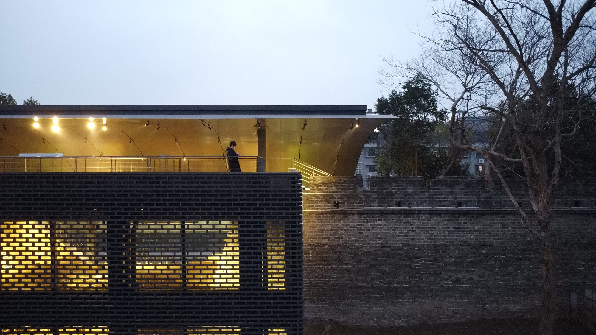 Nighttime view of the perforated brick facade with backlit openings and visitor on upper canopy level