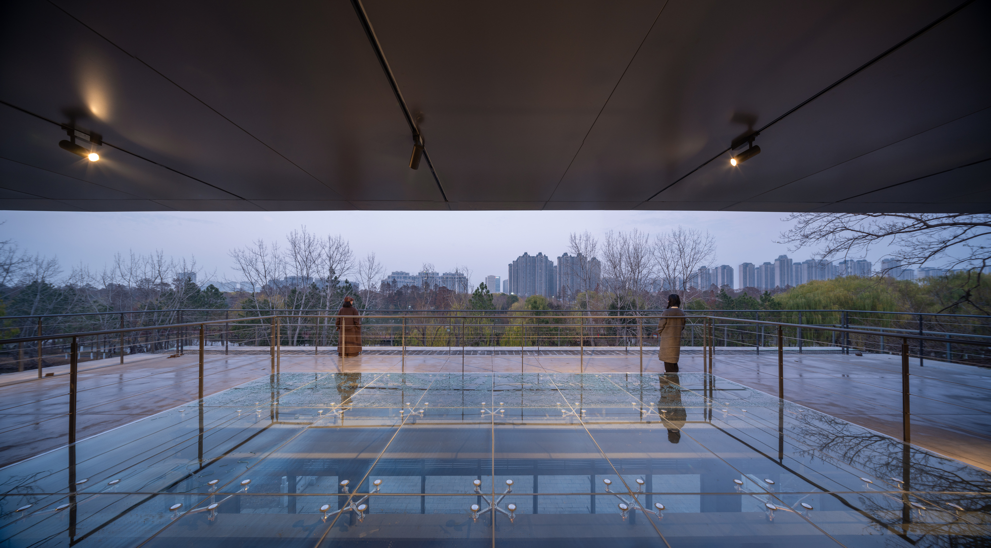 Glass floor terrace with steel railings reflecting bare trees and overcast sky