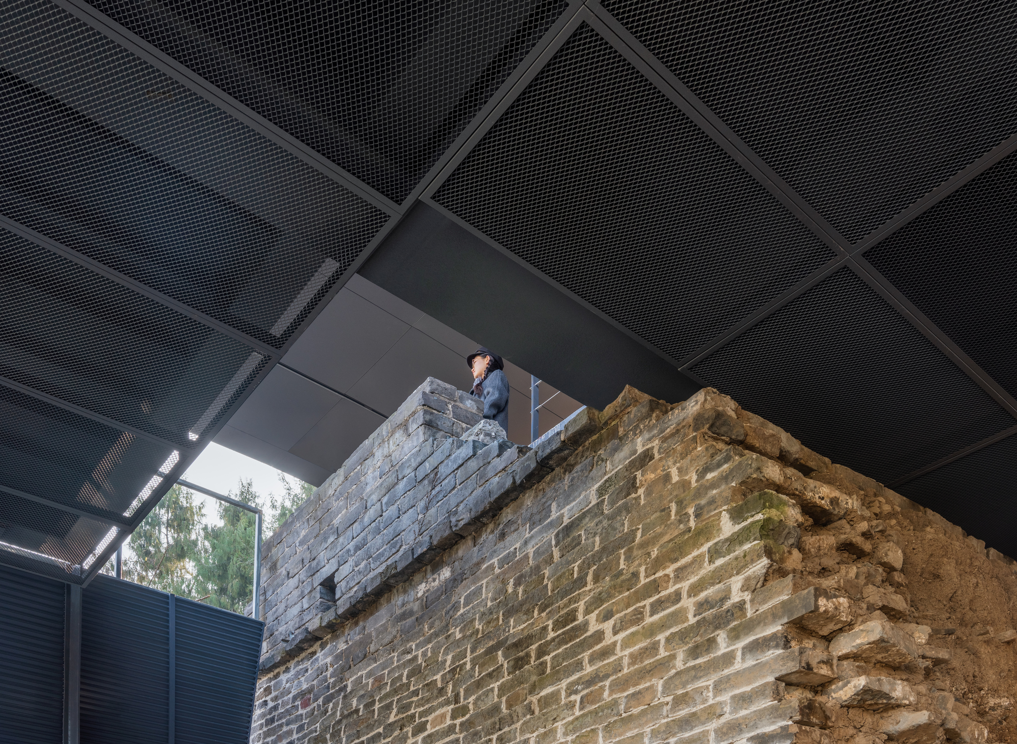 Visitor standing atop preserved brick wall remnant under perforated metal ceiling panels