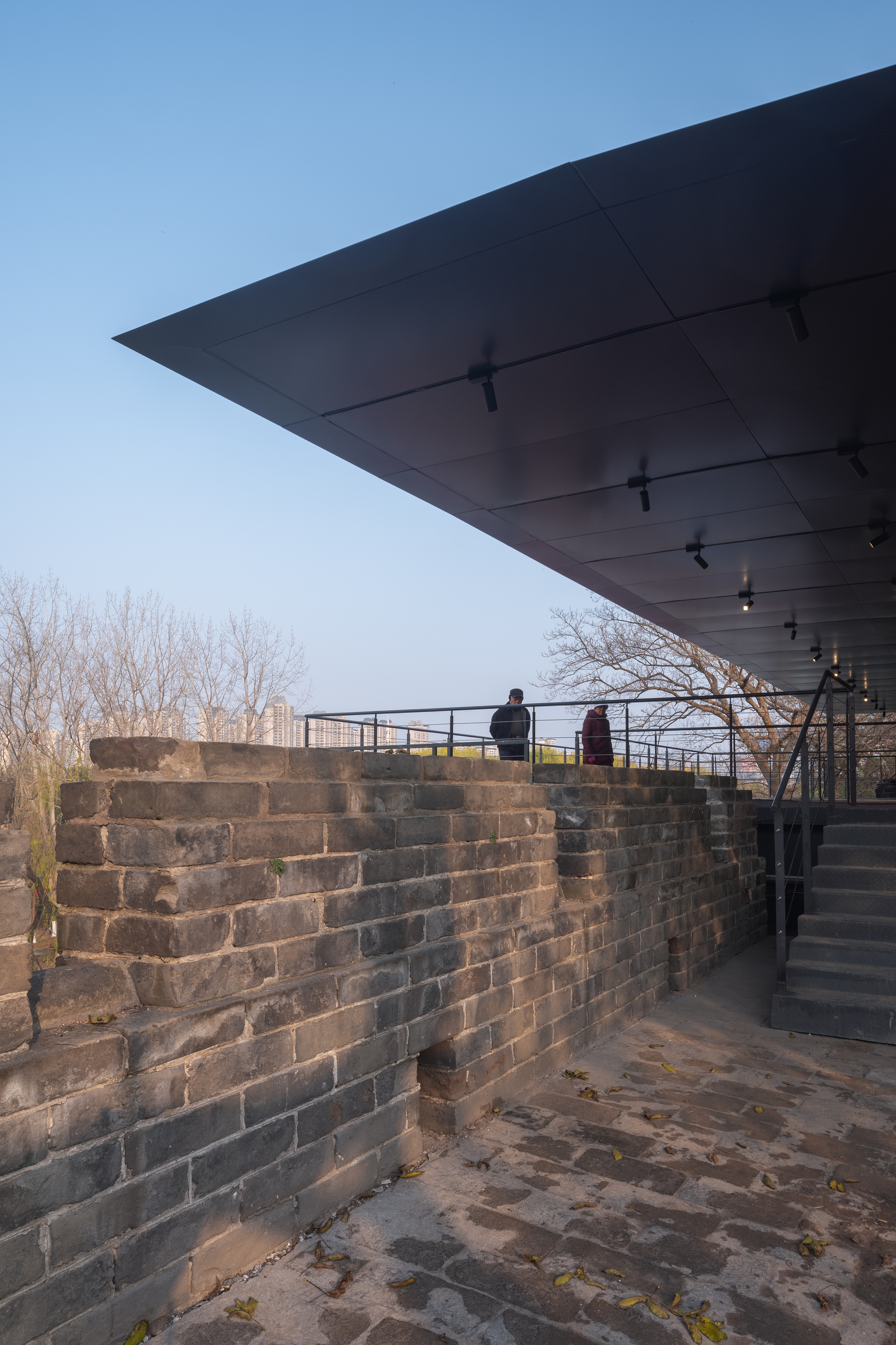 Cantilevered roof with track lighting extends over a historic brick wall path with two visitors at dusk