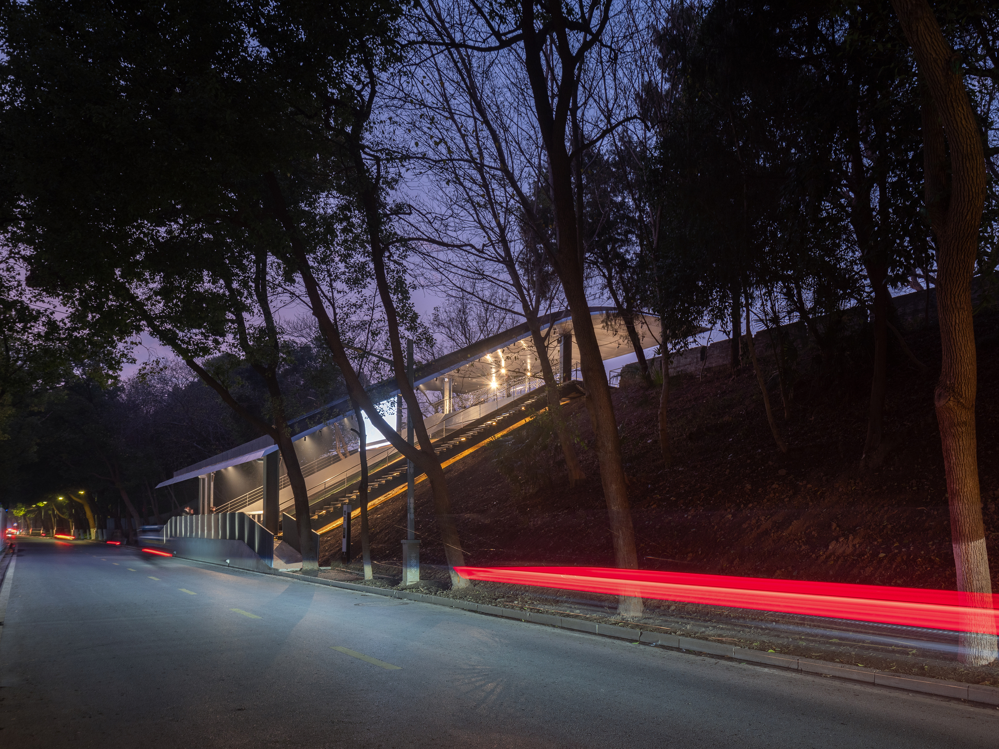 Street view of the illuminated structure against wooded hillside with car light trails passing at night