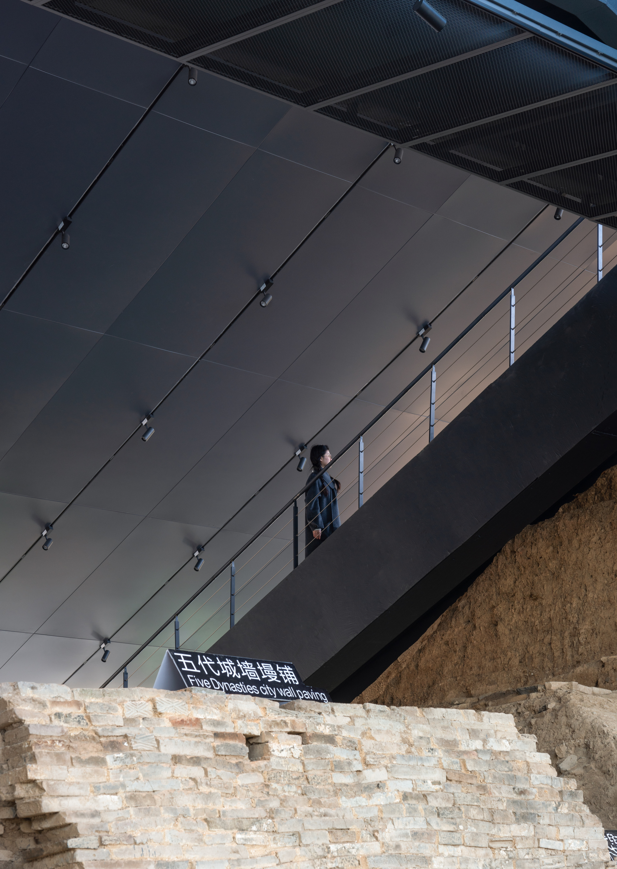 Person standing on the upper gallery above a reconstructed cream brick wall beneath a dark ceiling with track lighting