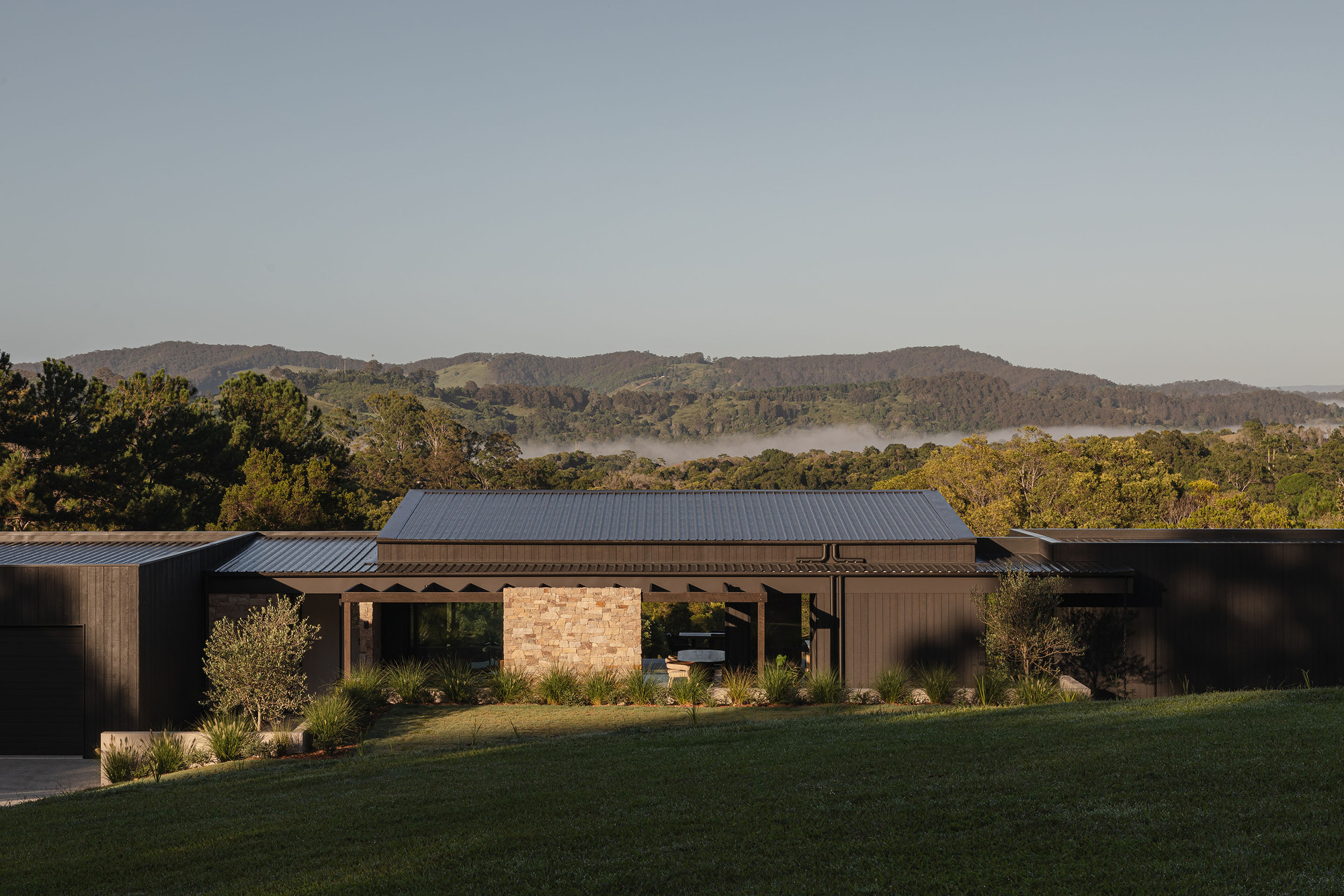 Front elevation with stone masonry core and vertical timber cladding set against forested hills in morning light