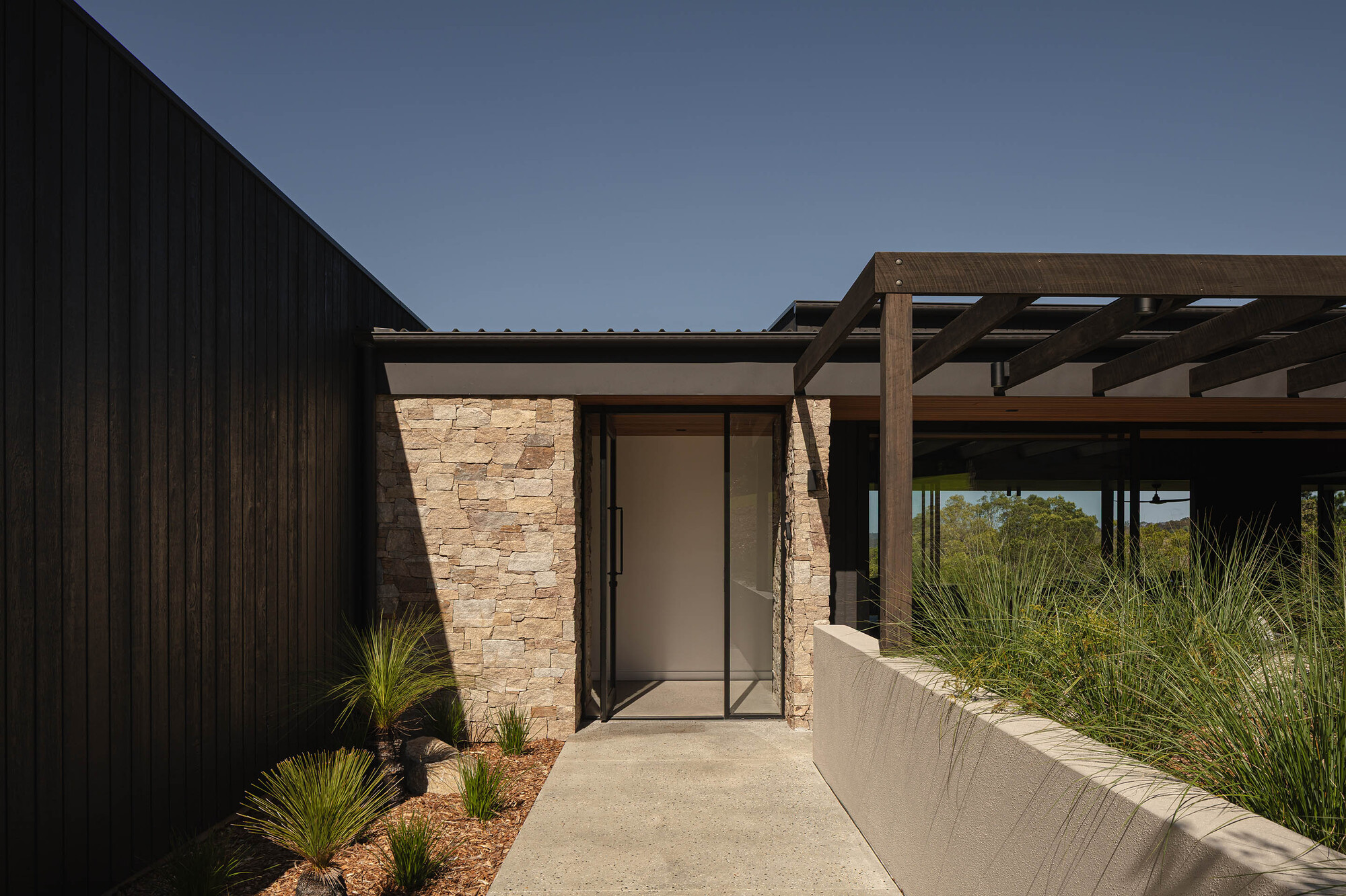 Entry pathway with stone veneer walls, dark vertical cladding, and timber pergola over planted beds