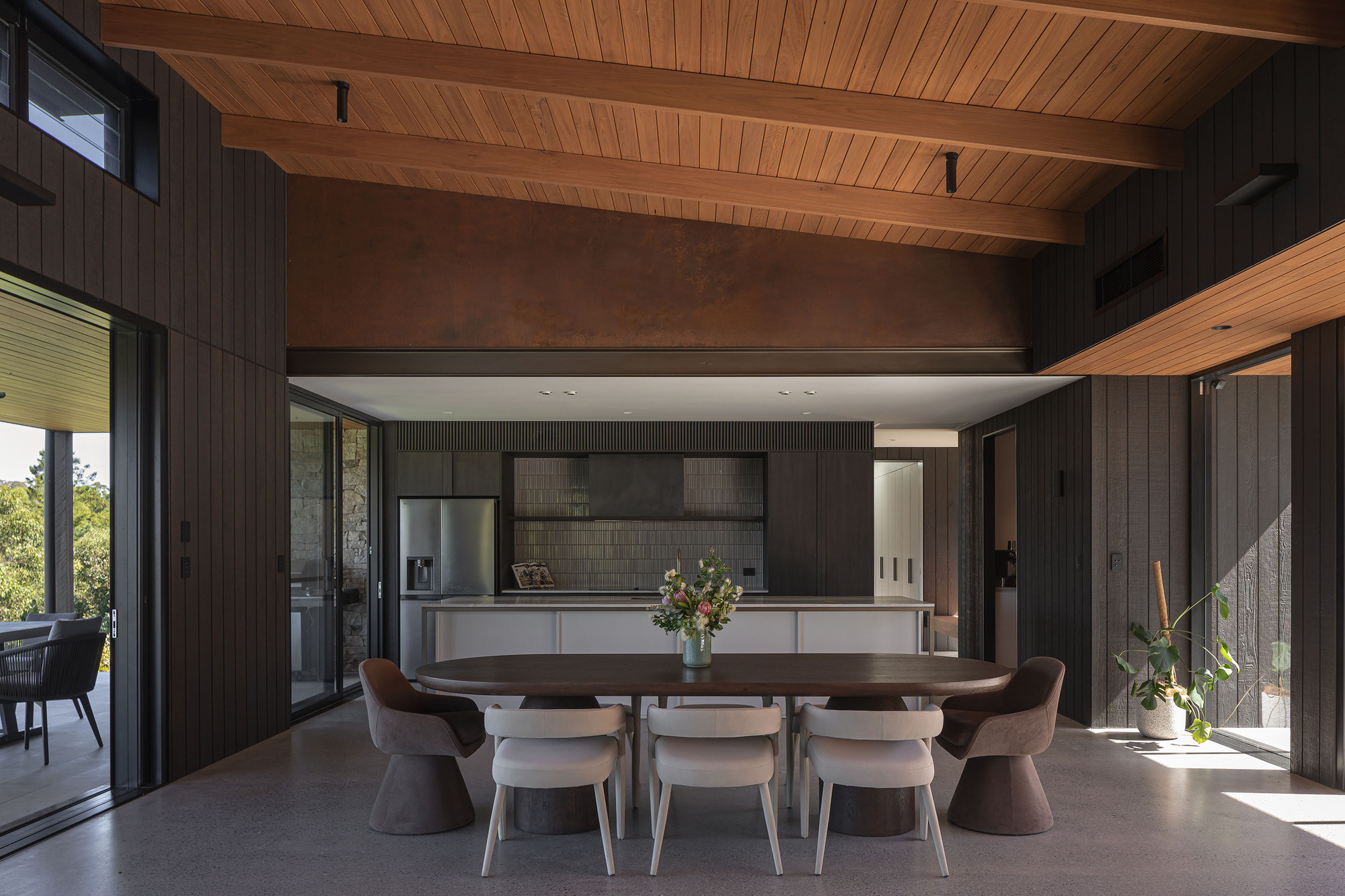 Open dining and kitchen area with timber ceiling beams and dark vertical wall cladding in afternoon light