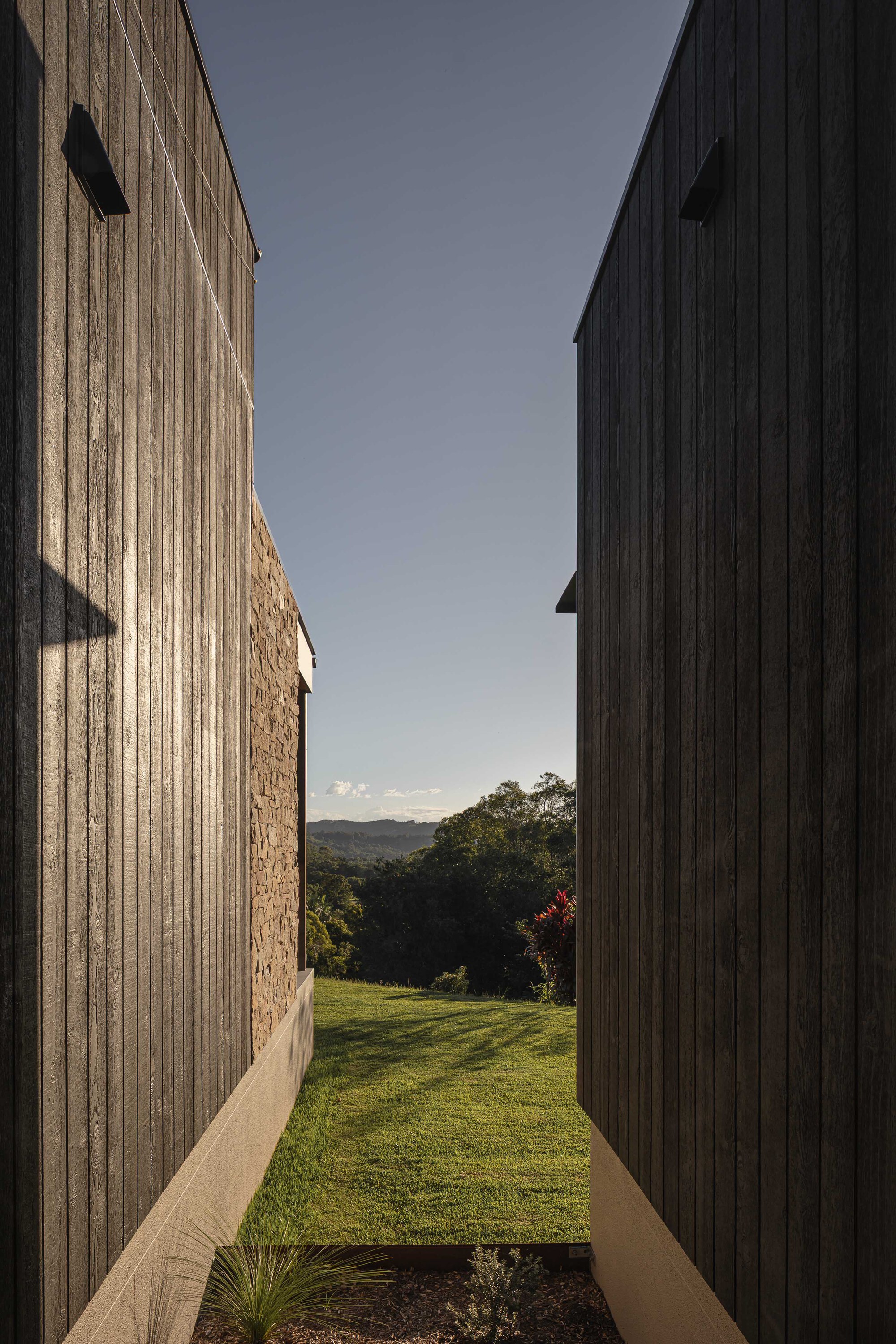 Narrow passage between vertical timber cladding and stone walls opening to lawn