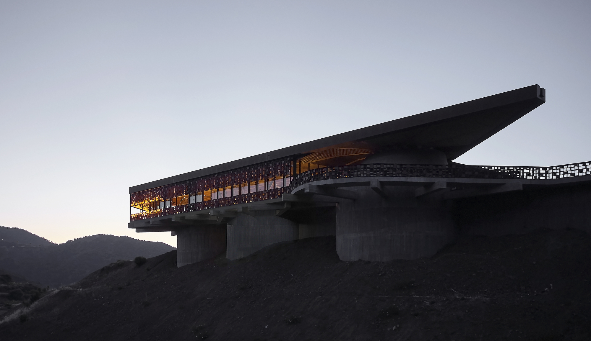 Cantilevered volume with mosaic screen facade and interior lighting at dusk above concrete piers