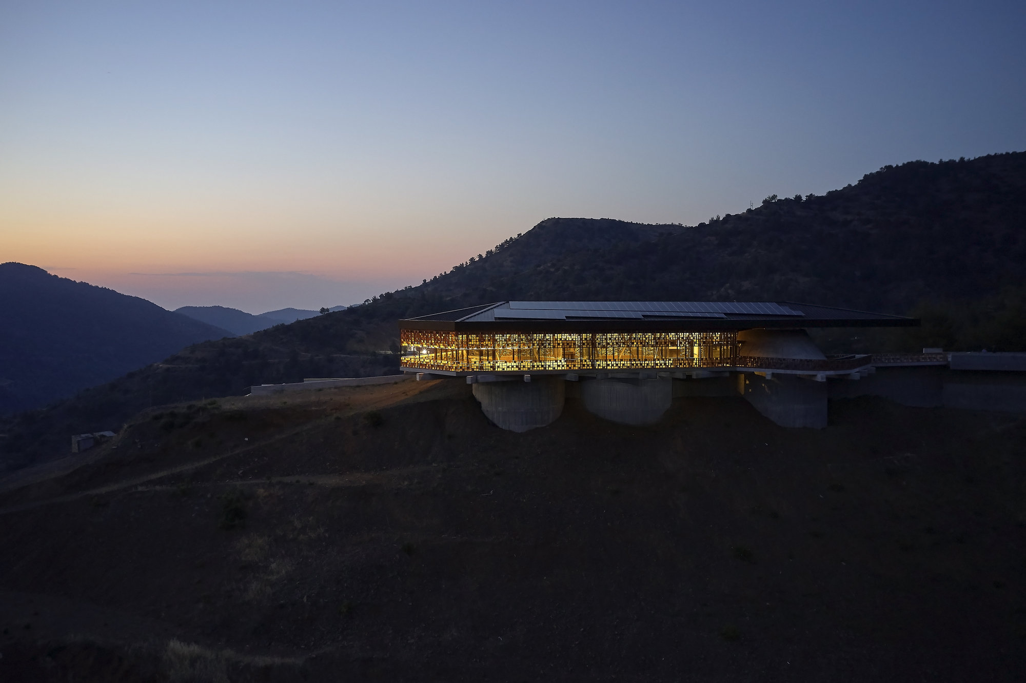 Illuminated glass facade perched on concrete piers against forested hills at dusk