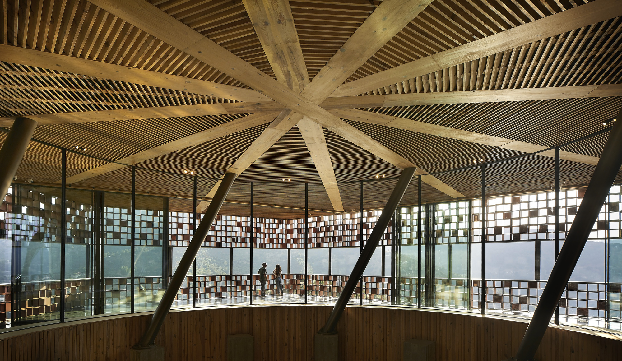 Interior view of radial timber beams converging at the ceiling center above glass-walled galleries with visitors