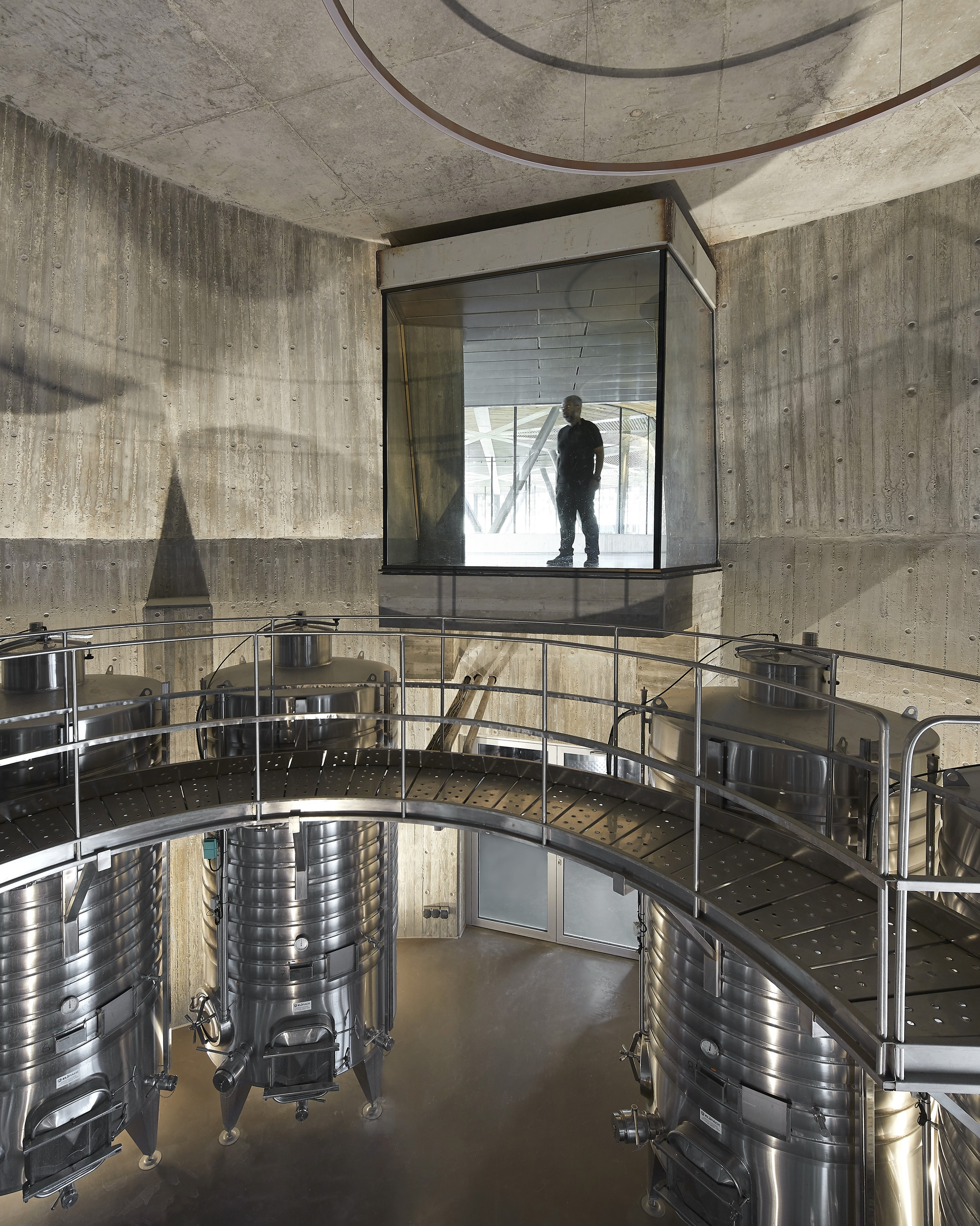 Glass viewing box cantilevered over production floor with stainless steel fermentation tanks and board-formed concrete walls