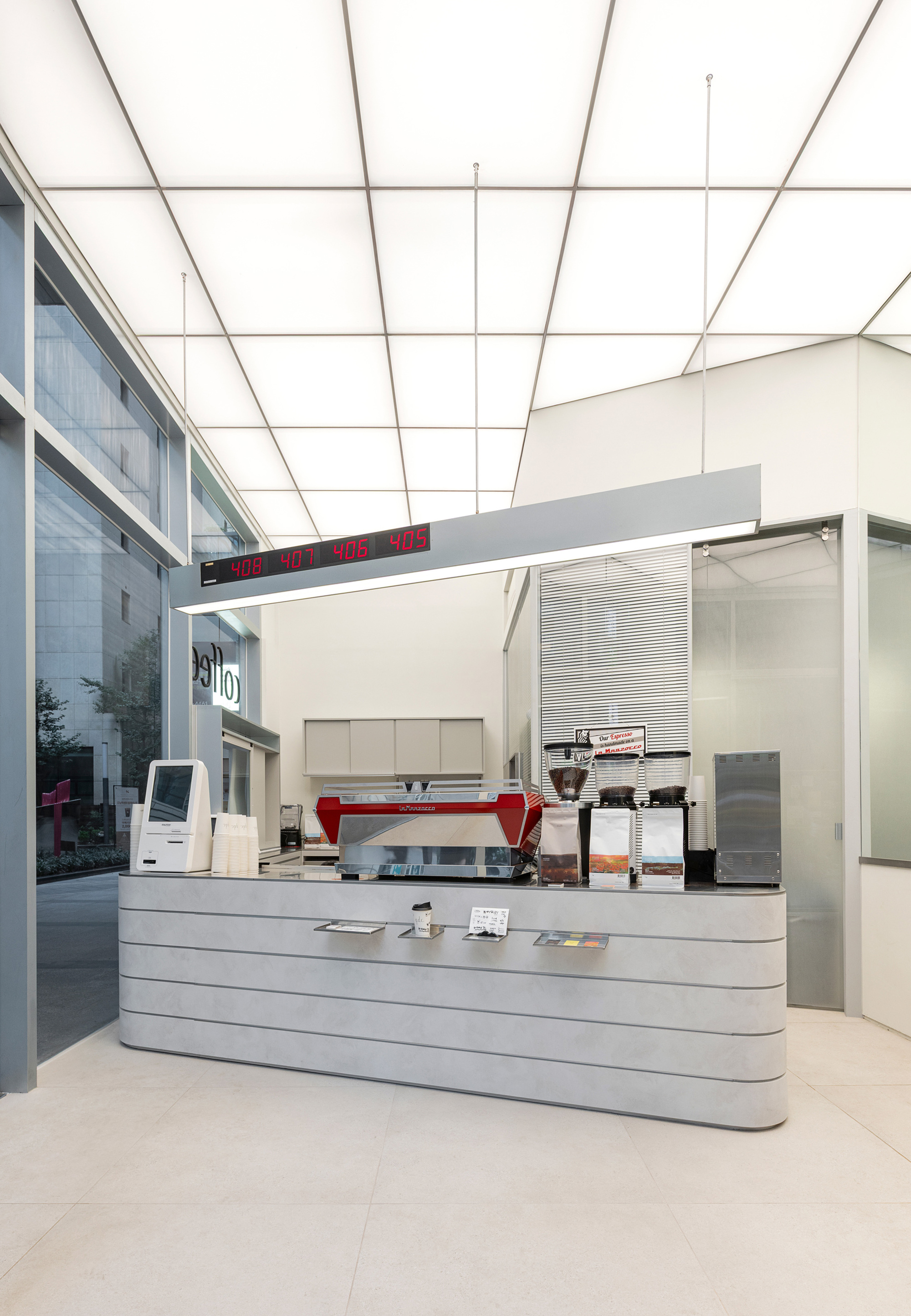 Red espresso machine on ribbed counter beneath translucent panel ceiling and glazed courtyard