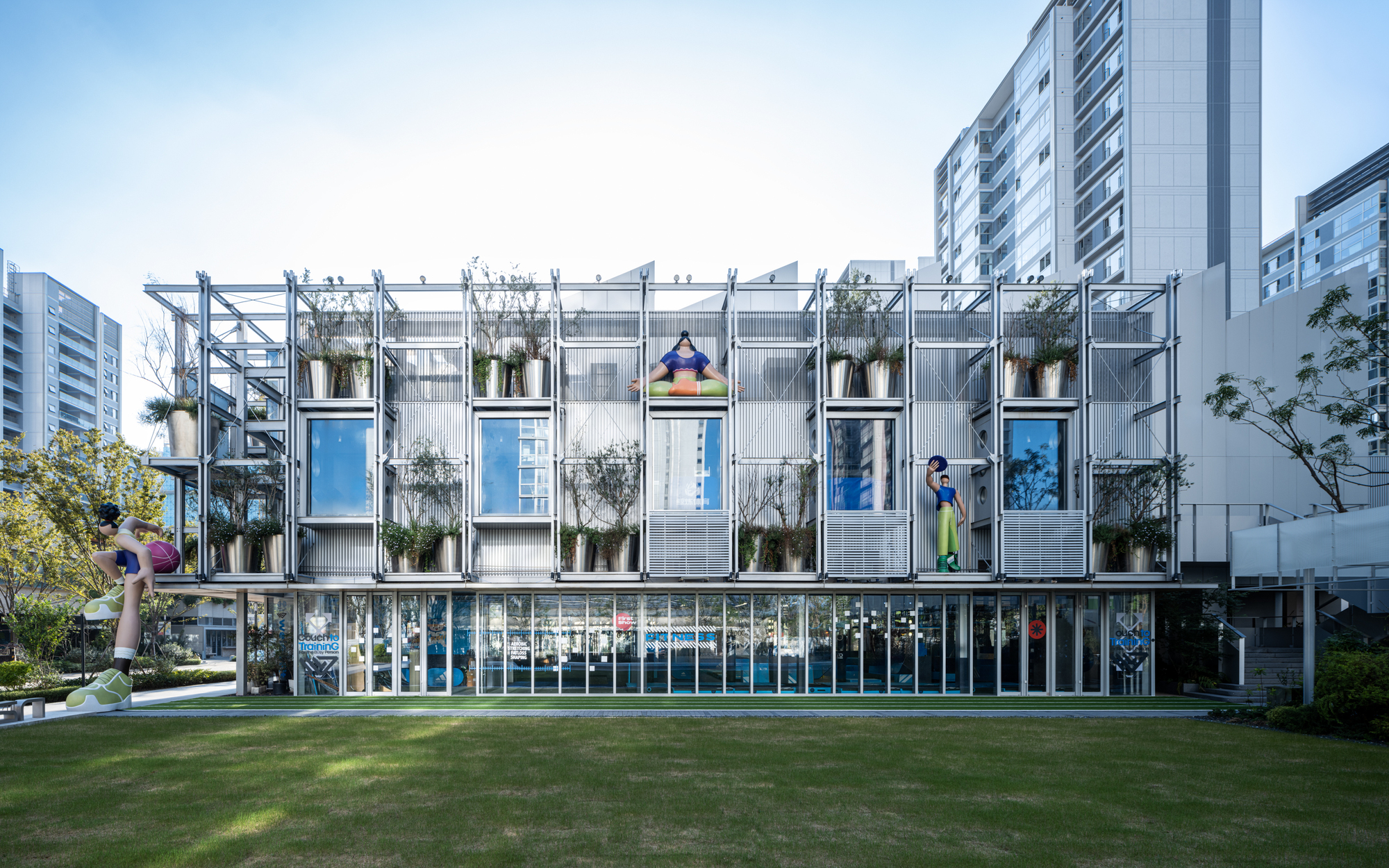 Front elevation of steel-framed structure with modular units and hanging plants viewed across a sunlit lawn