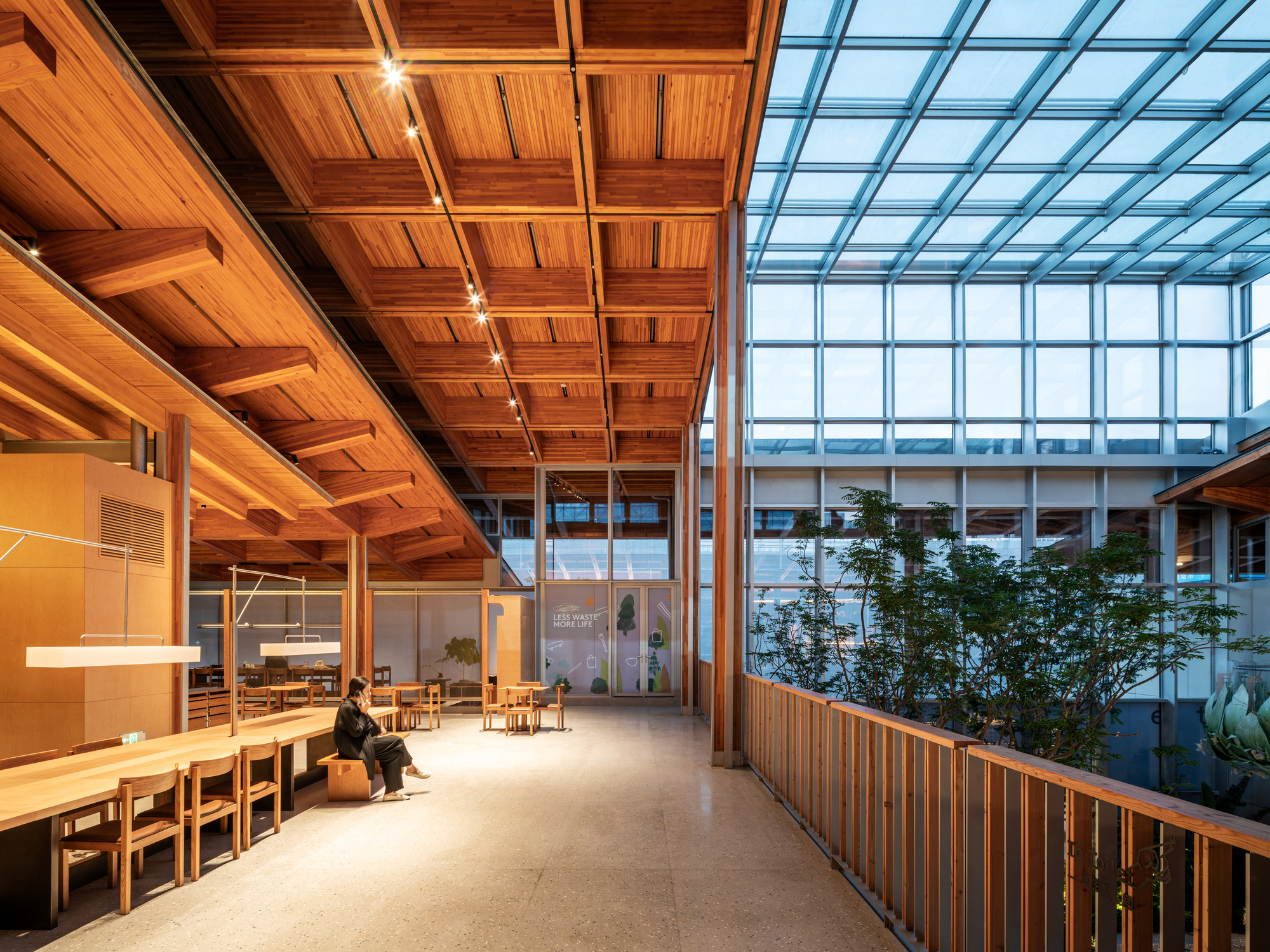 Interior corridor with coffered timber ceiling and central glass atrium with planted trees at dusk