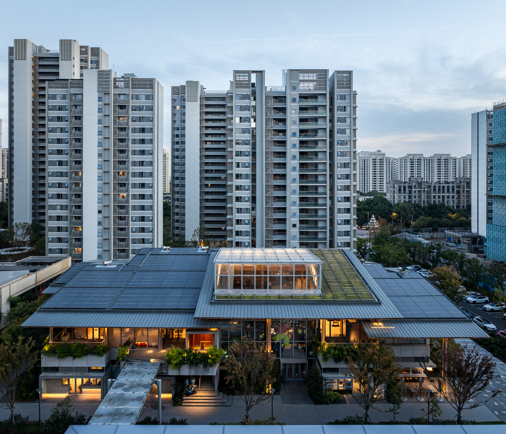 Aerial view of low-rise timber structure with roof terrace surrounded by residential towers at dusk