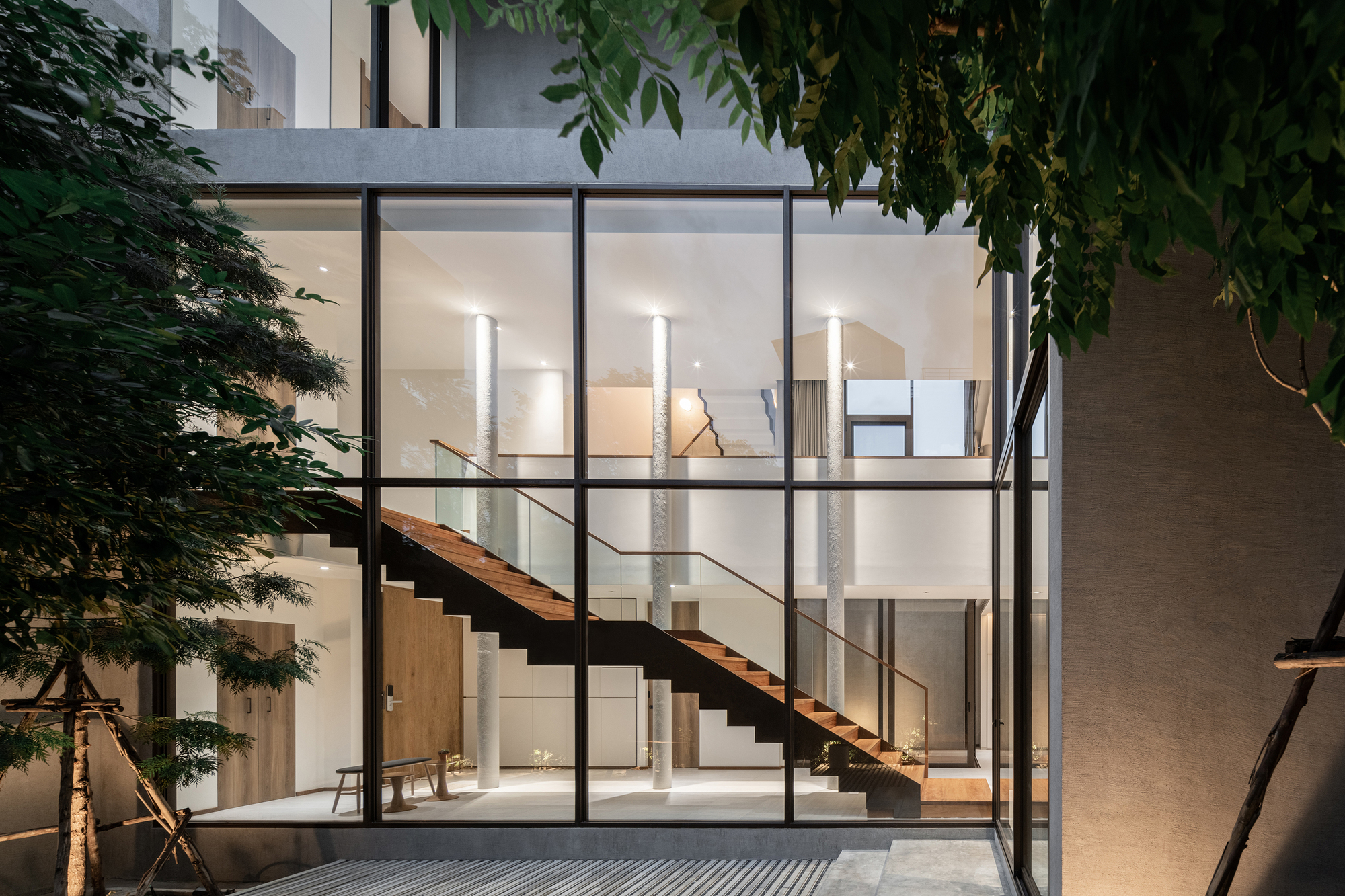 Courtyard view at twilight showing illuminated staircase through gridded glass facade surrounded by trees
