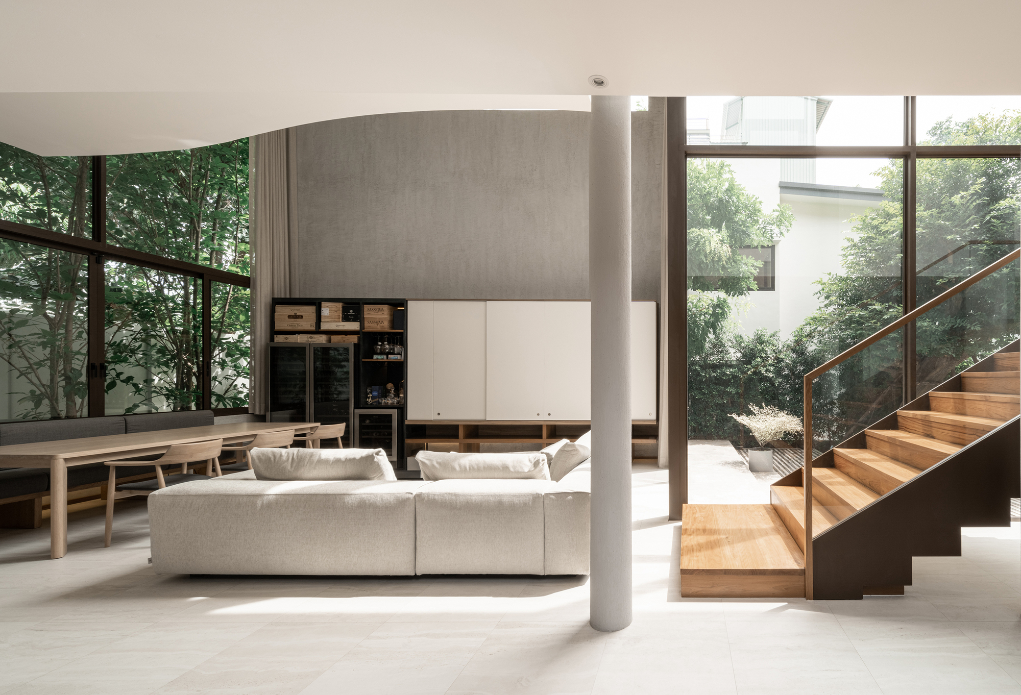 Living area with timber staircase, cylindrical columns, and floor-to-ceiling glazing overlooking greenery