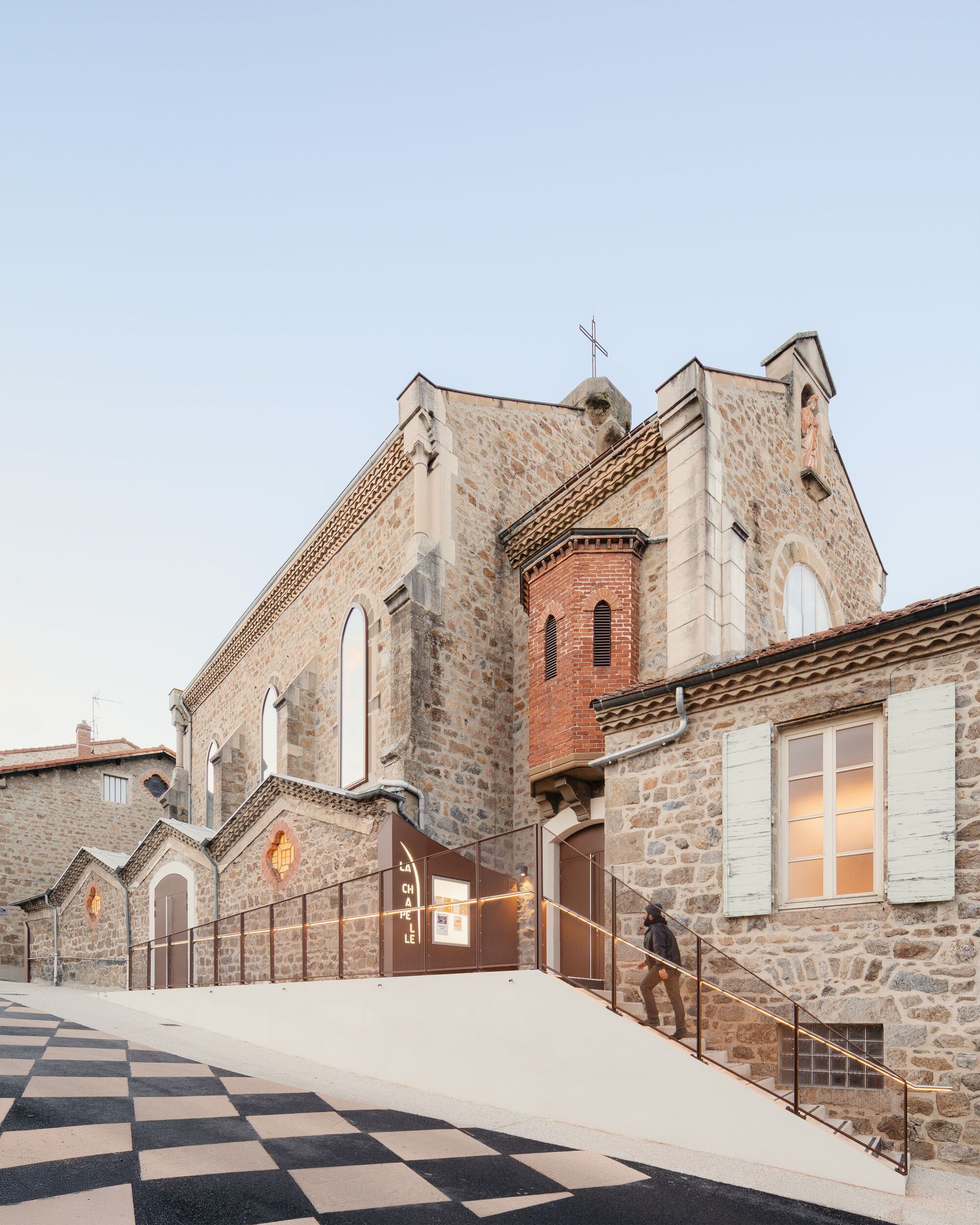 Exterior courtyard with chequered paving leading to illuminated church entrance at twilight