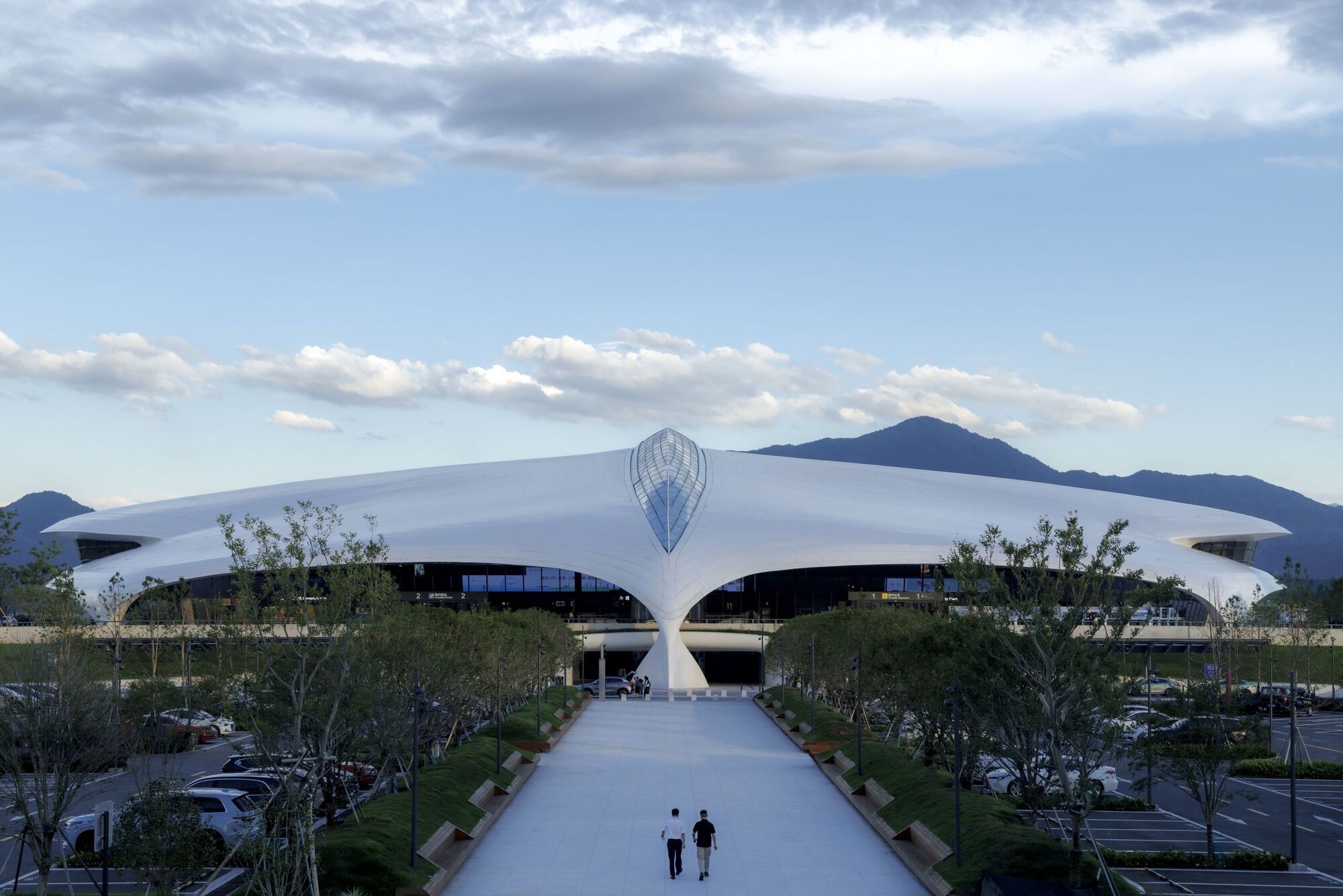 Two visitors walking the central arrival pathway toward the white curving roof canopy with mountains in the background
