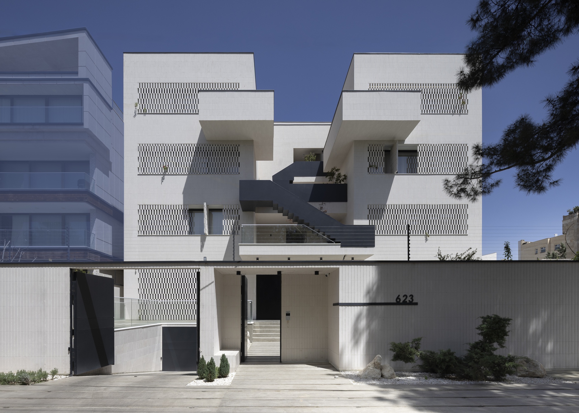 Street facade with stacked balconies and perforated white screens behind a concrete wall with planted entry court