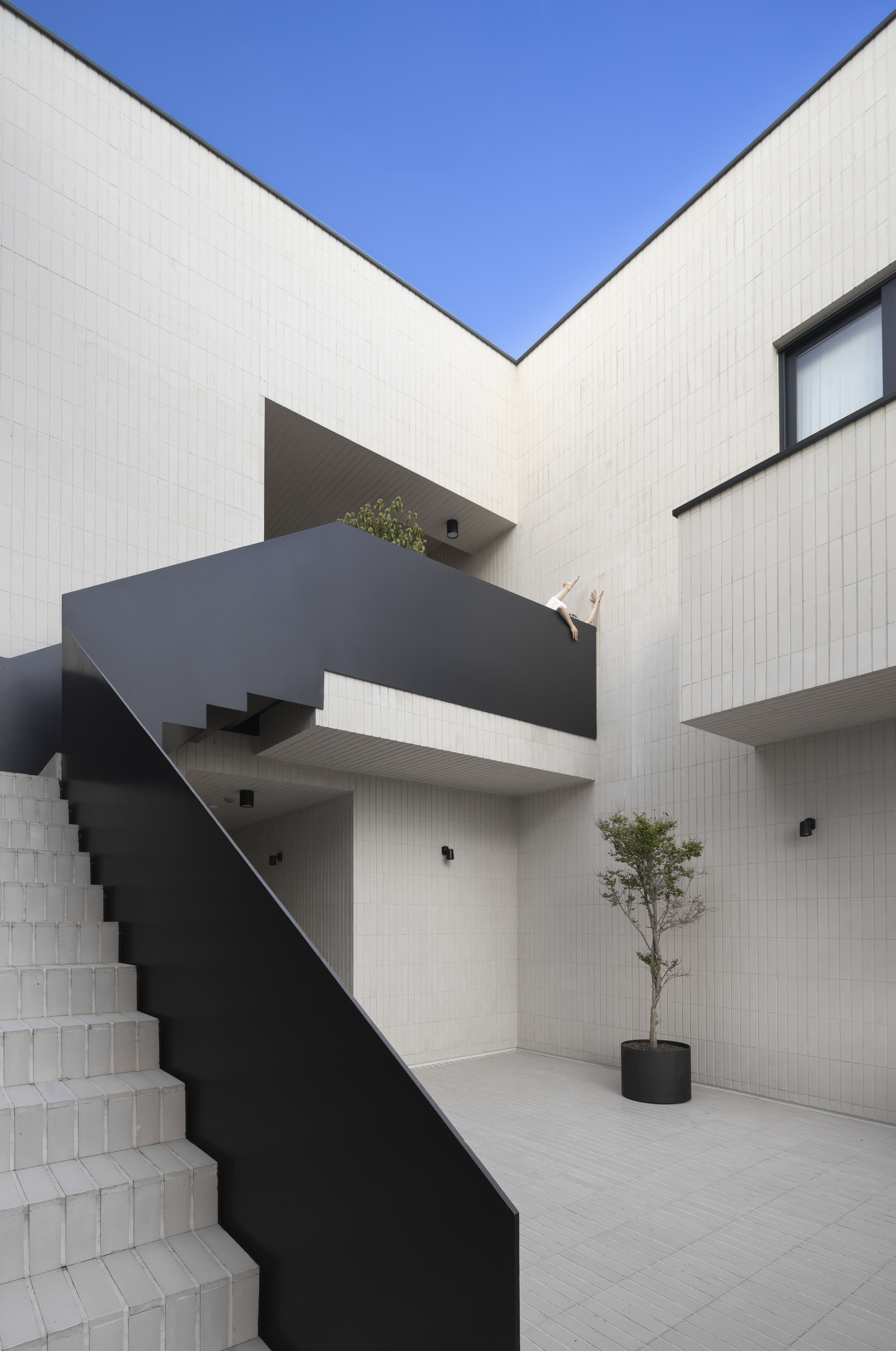 Internal courtyard with folded black steel staircase and planted bed against white tiled walls under clear sky