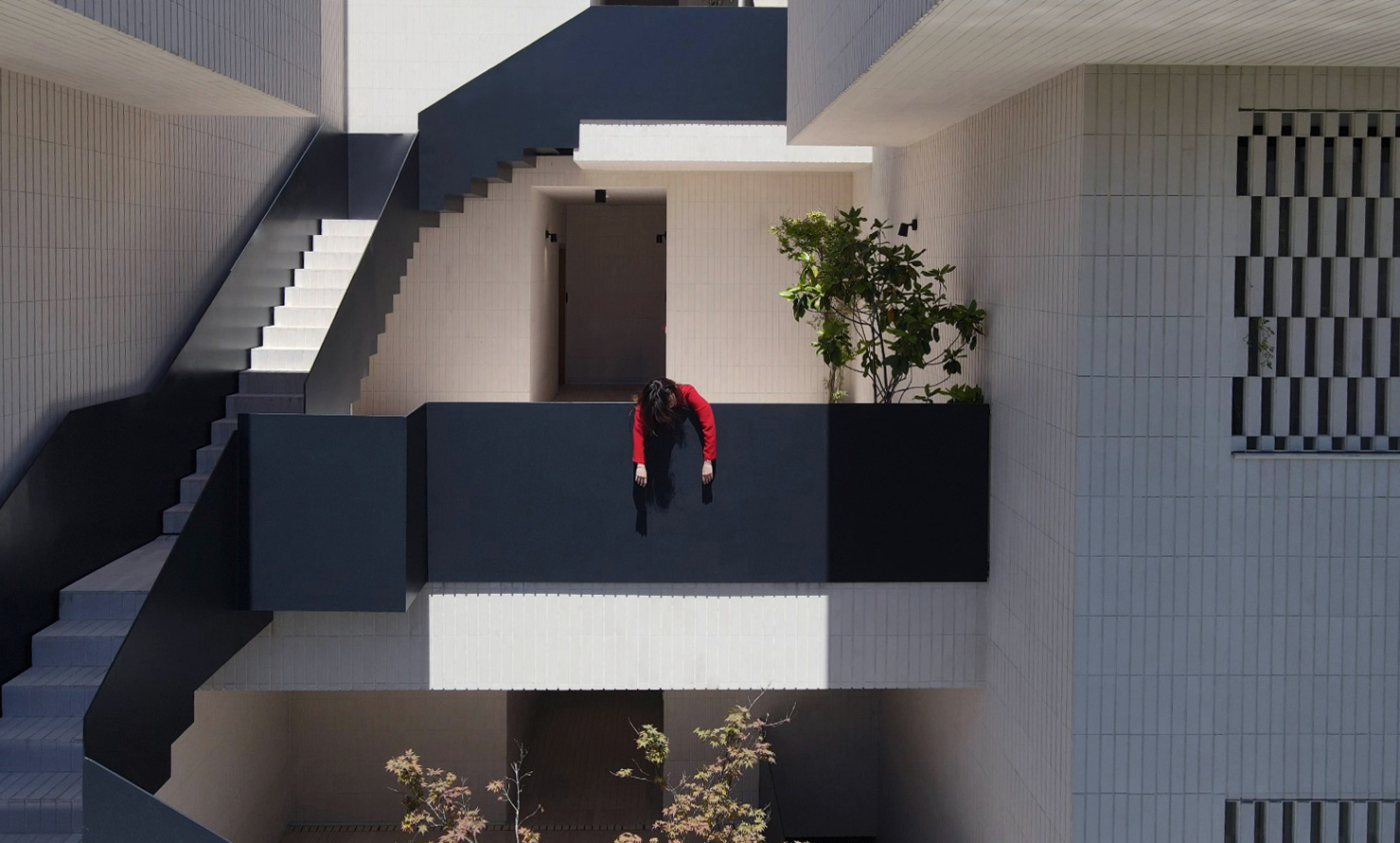 Overhead view of a figure in red leaning over the black steel railing of a courtyard bridge