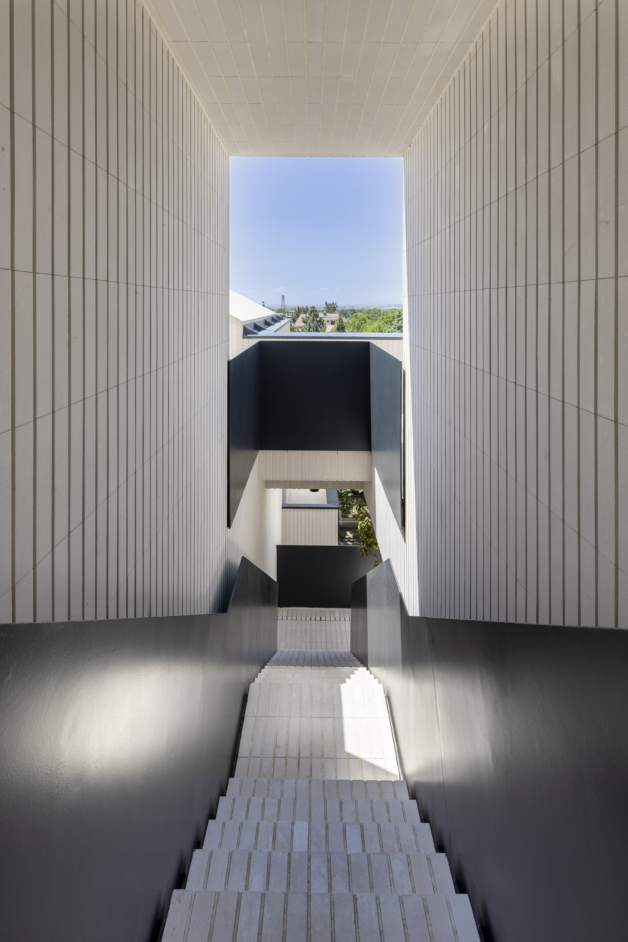 Vertical stairwell framed by white ribbed walls rising toward a skylight opening above black steel landings
