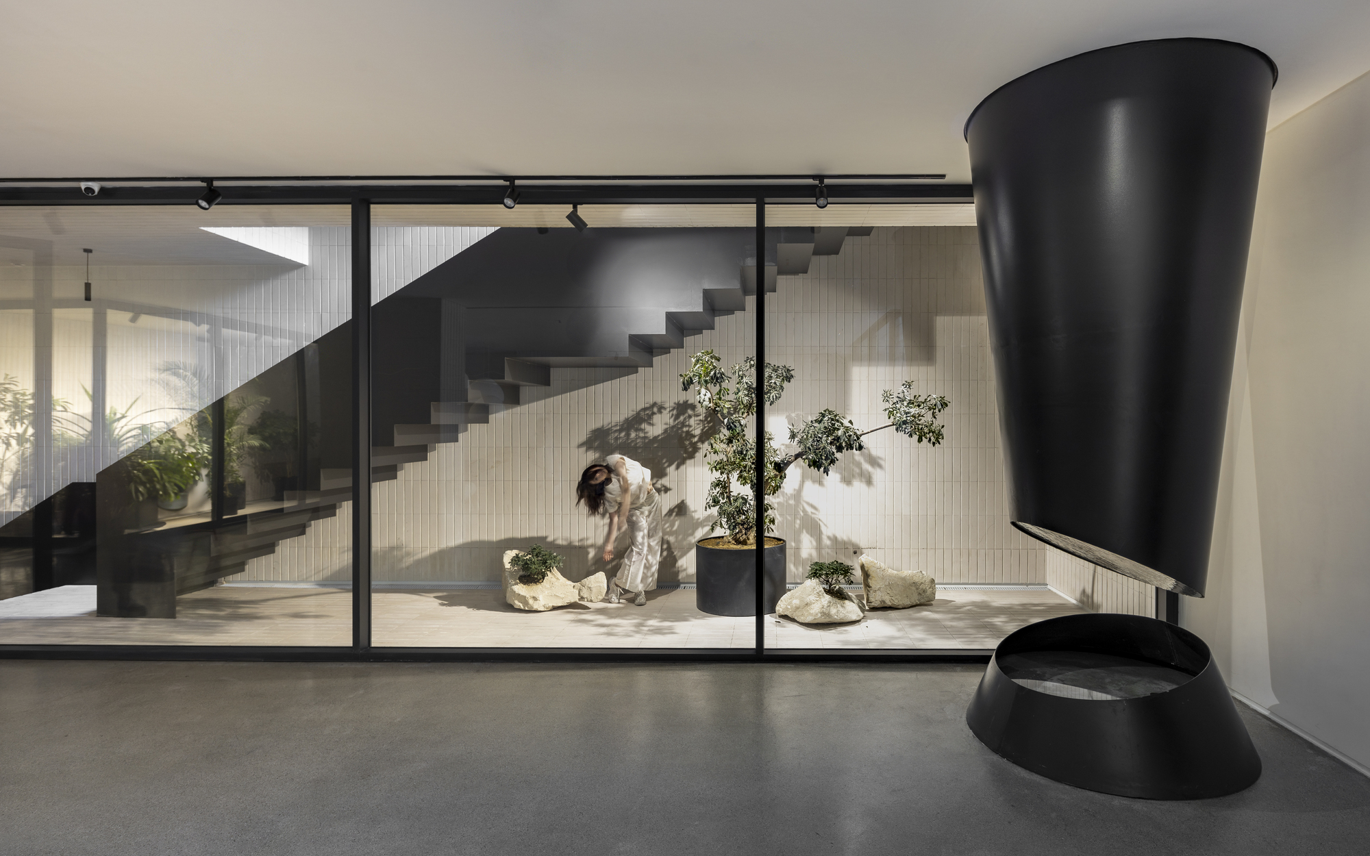 Interior view through glazed partition showing cantilevered black staircase and person tending bonsai plants