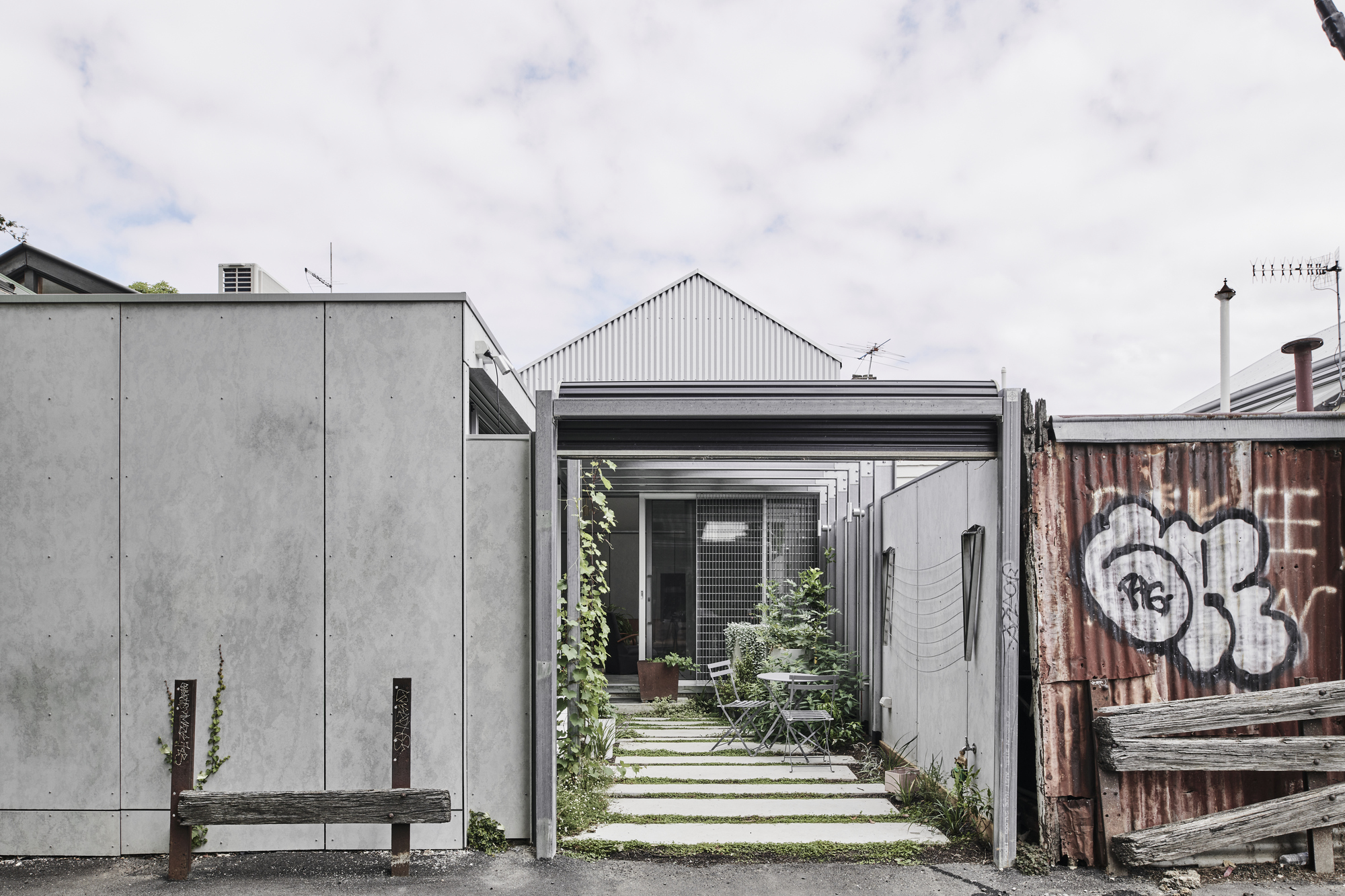 Narrow laneway entrance framed by concrete panels and corrugated metal shed with weathered timber fence