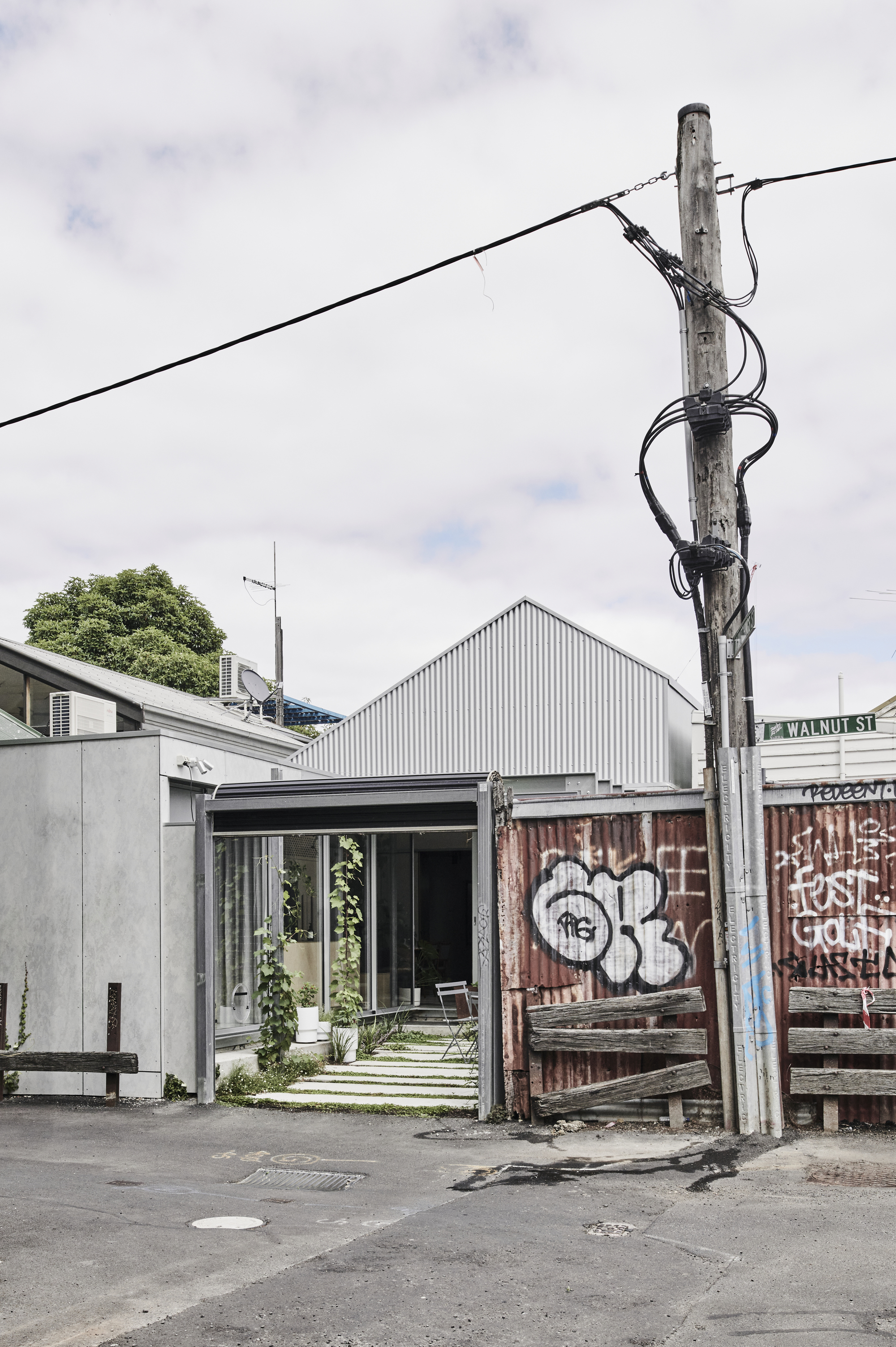 Street view of corrugated metal gable roof volume beside graffitied timber fence and utility pole