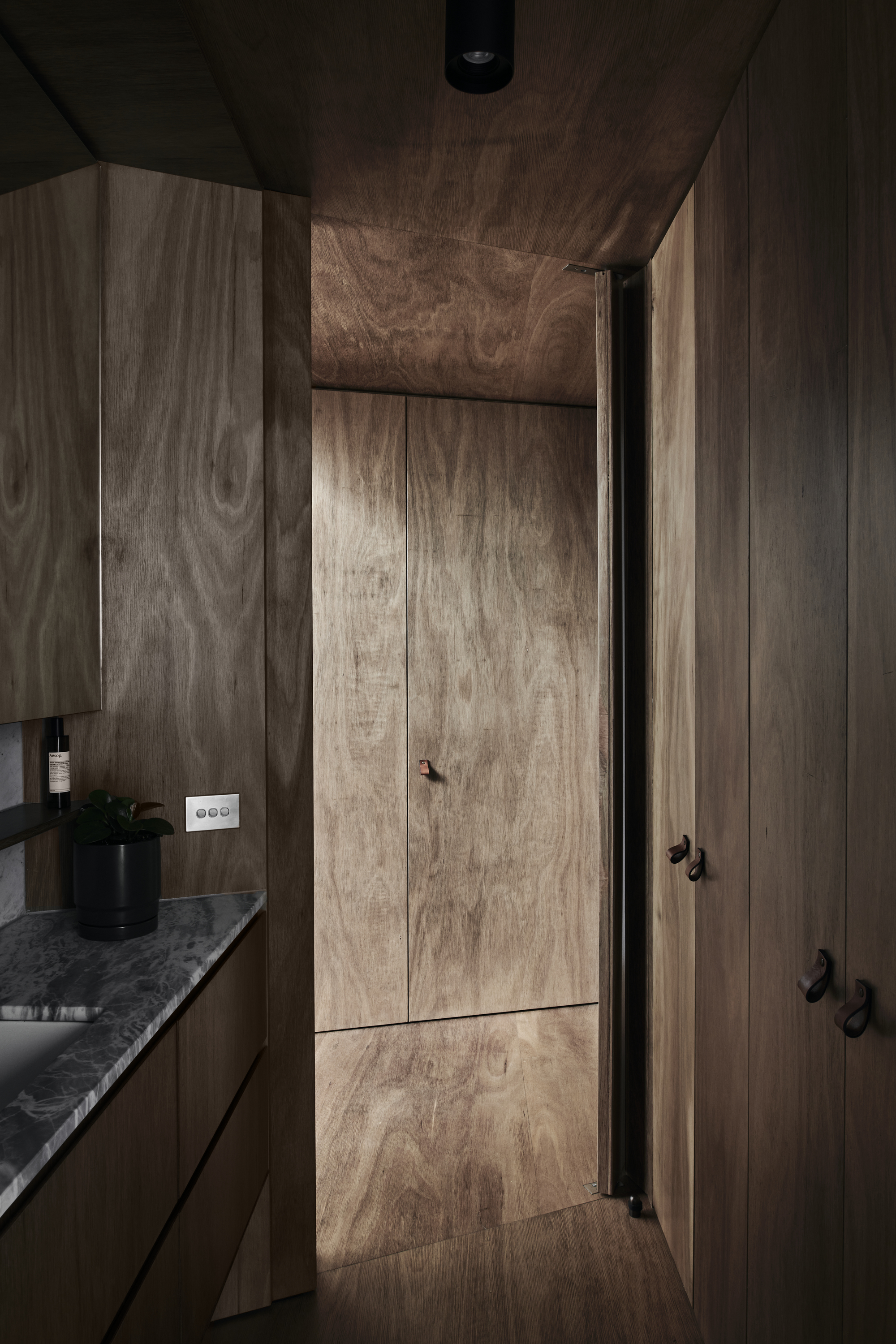 Narrow corridor clad entirely in timber veneer with flush doors and a stone countertop glimpsed beyond