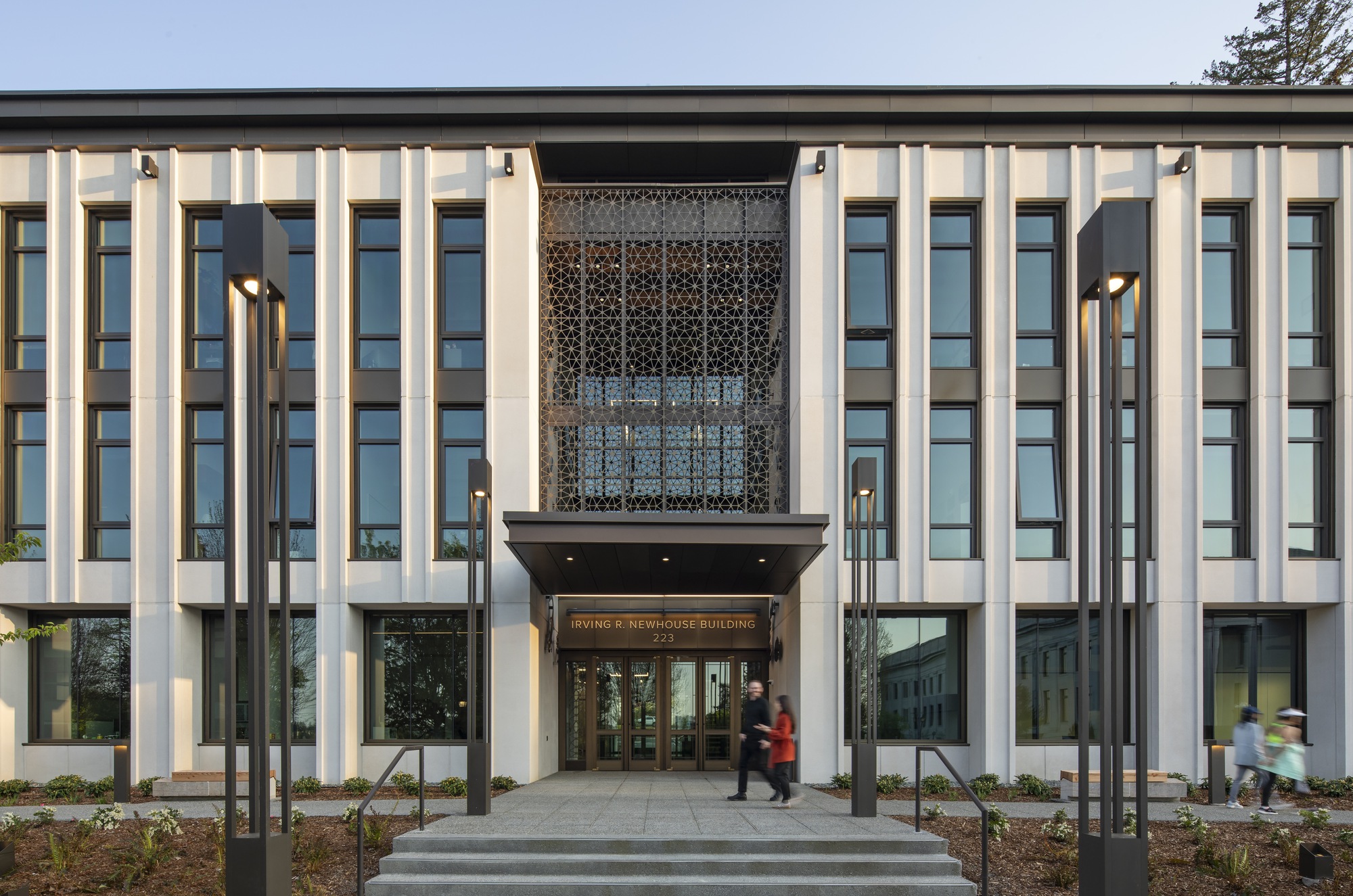 Entry facade showing vertical fins flanking a perforated metal screen above glazed doors with people entering at twilight