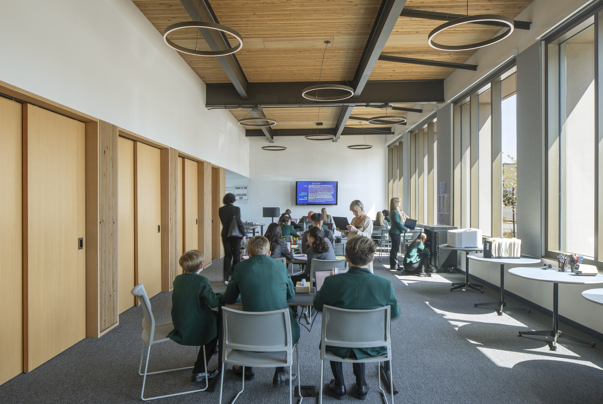 Interior study space with exposed steel beams, timber ceiling, circular pendant lights, and students gathered at tables