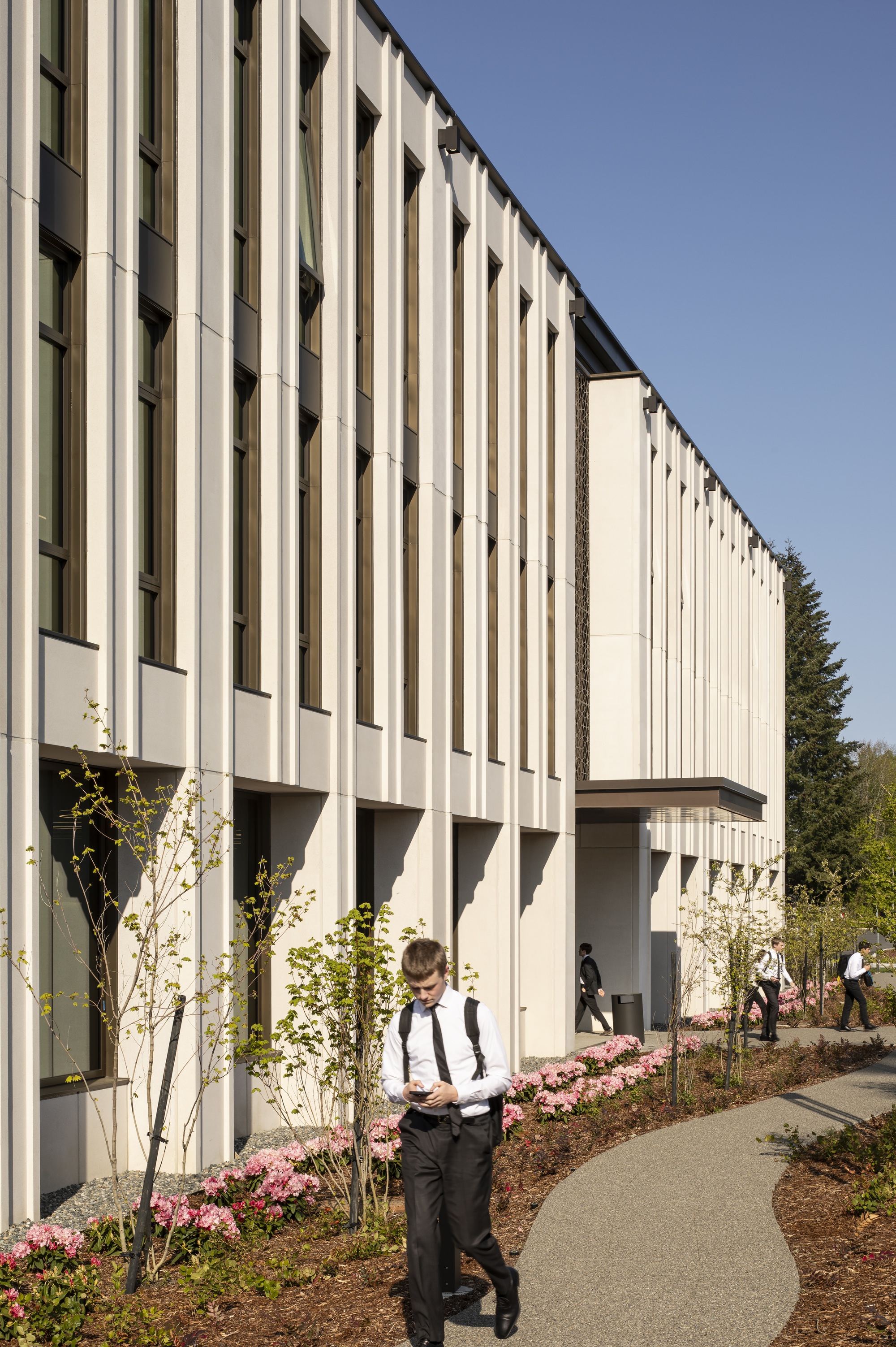 Side facade with vertical white fins and planted beds along the curving walkway in afternoon sunlight