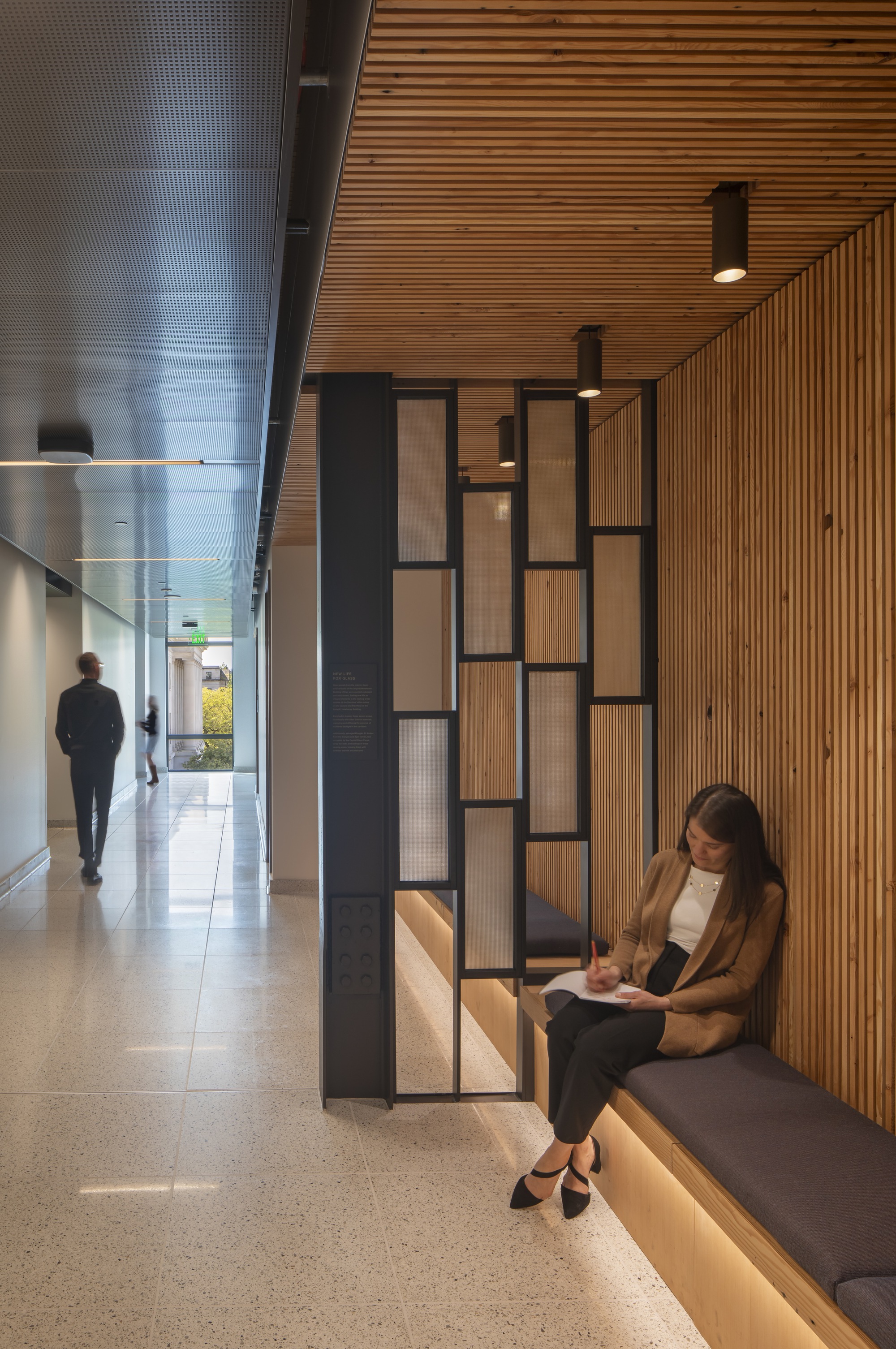 Corridor with slatted timber ceiling and geometric seating nook where a person reads