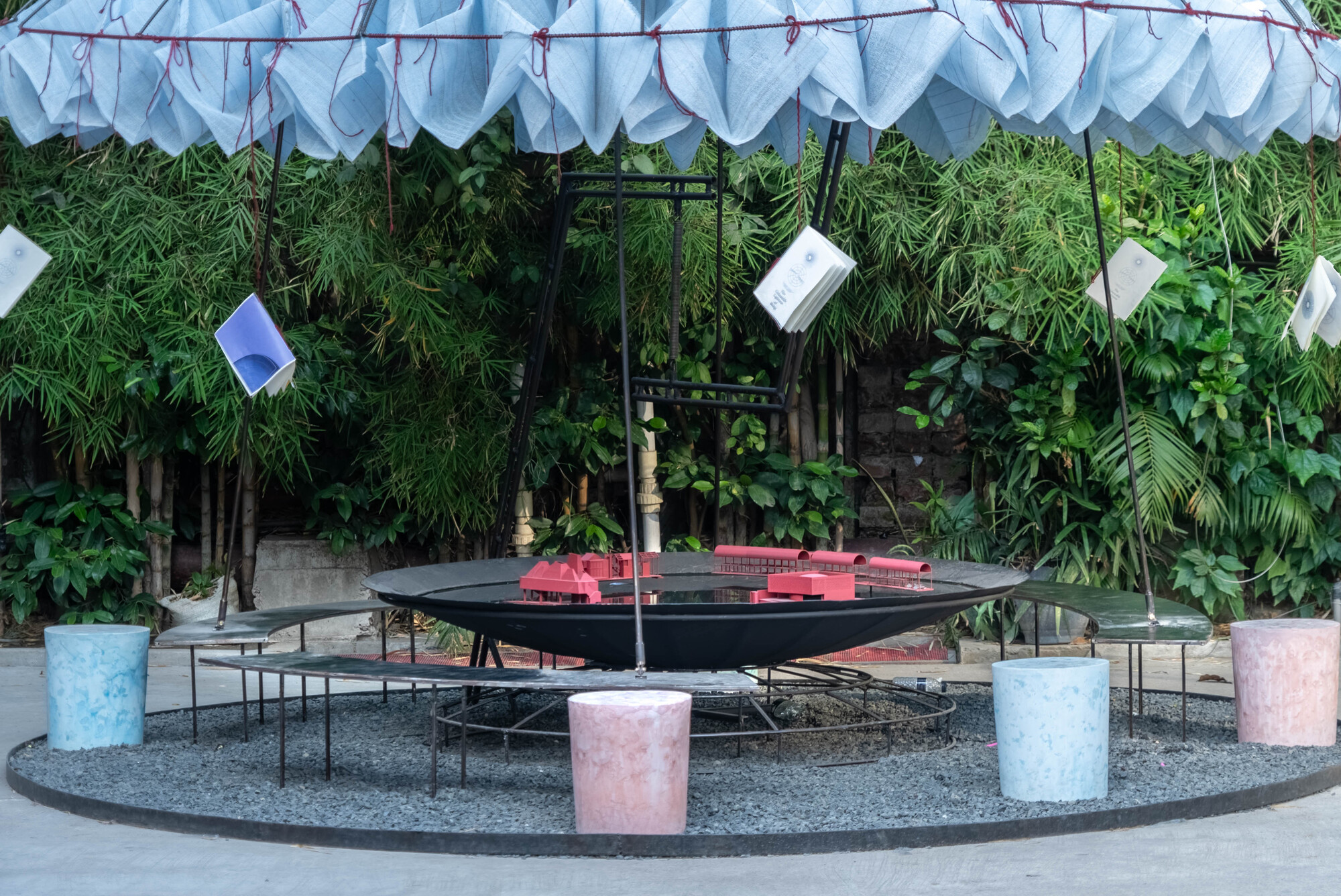 Circular installation with black metal table displaying red model buildings beneath a white fabric canopy and bamboo