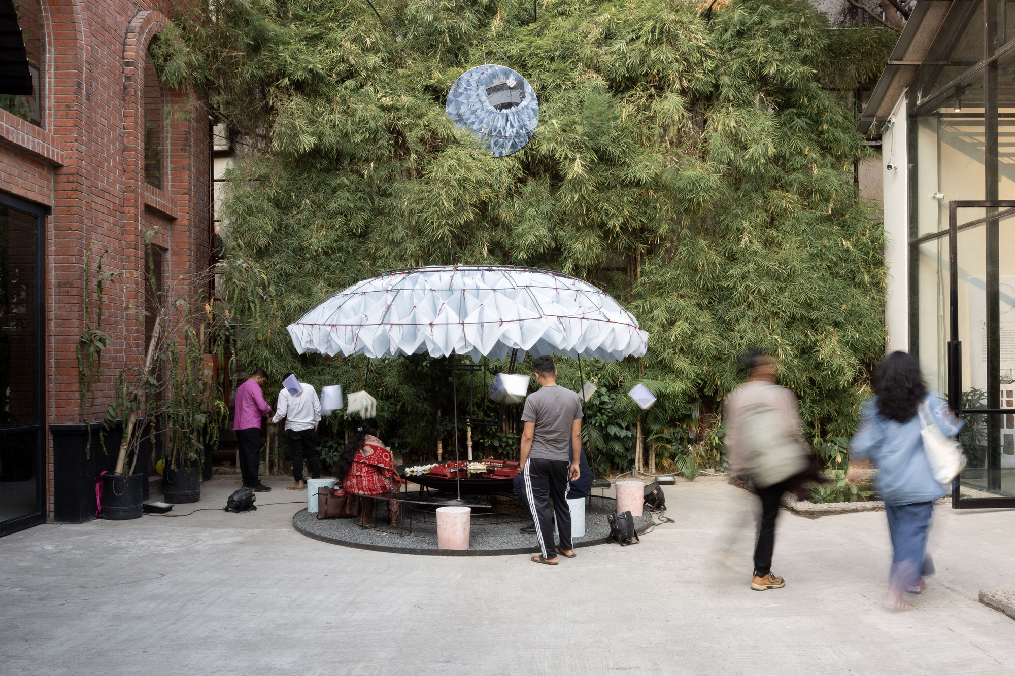 Courtyard installation with white canopy structure as visitors gather and walk past planted bamboo and brick facade