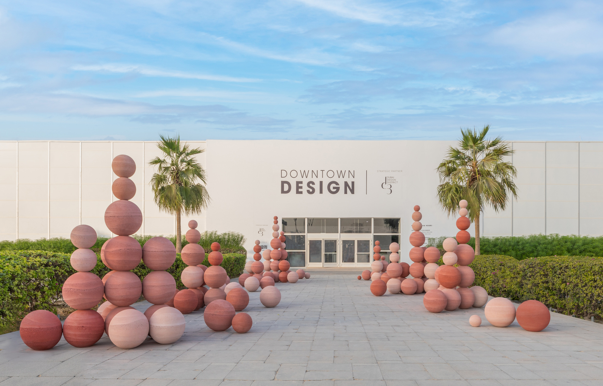 Stacked terracotta sphere installation on a paved plaza flanked by palm trees and hedges