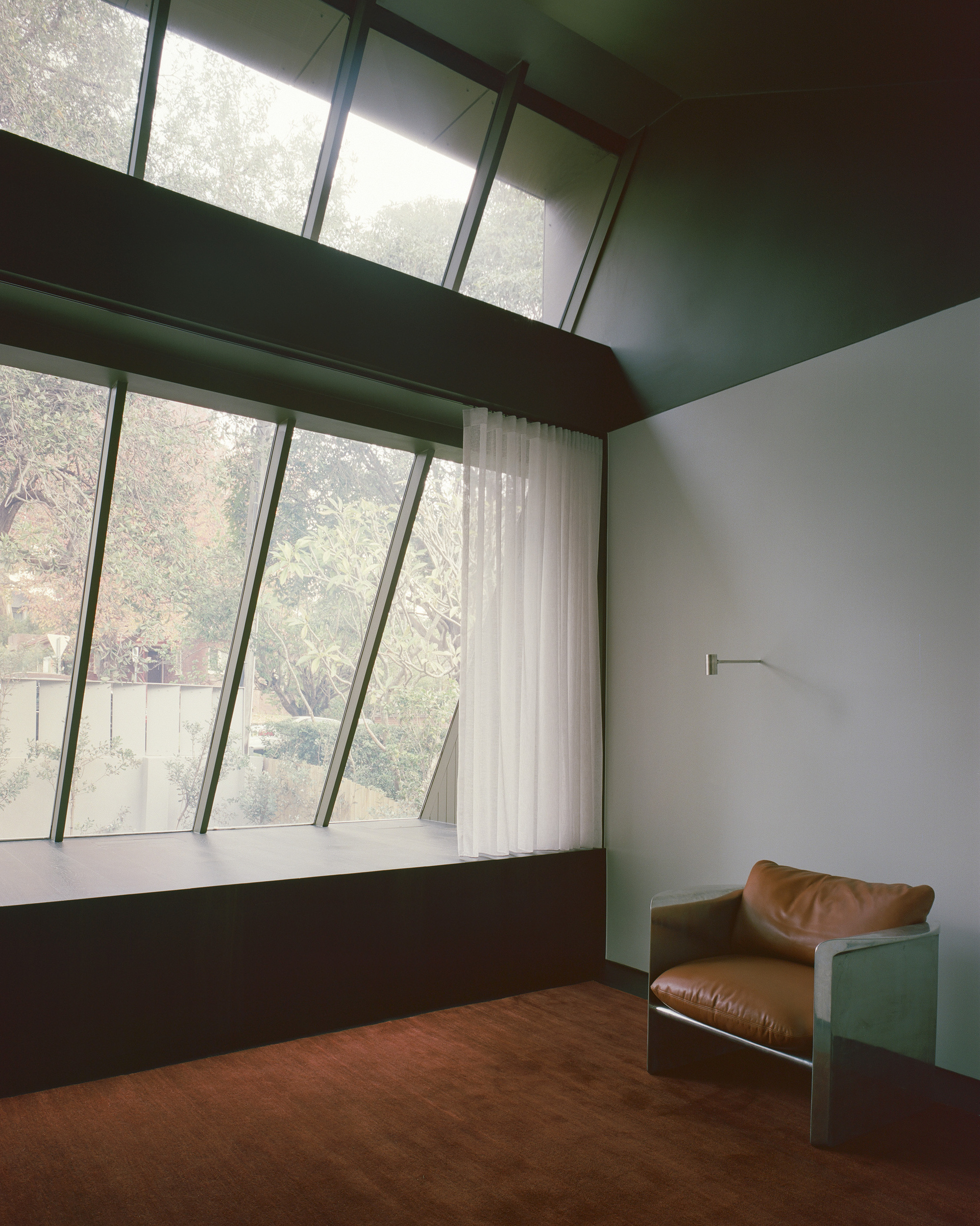 Bedroom with angled windows and timber beams overlooking garden vegetation through sheer curtains