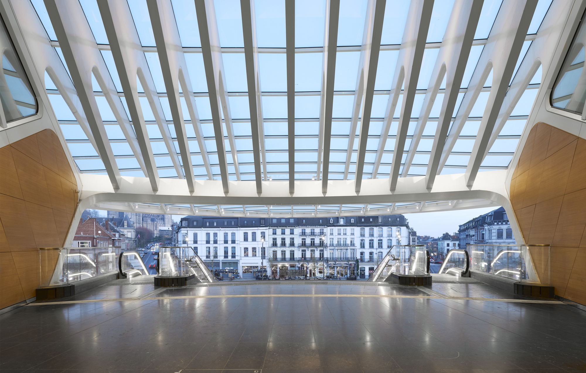 Station concourse with white steel and glass canopy framing historic buildings beyond escalators