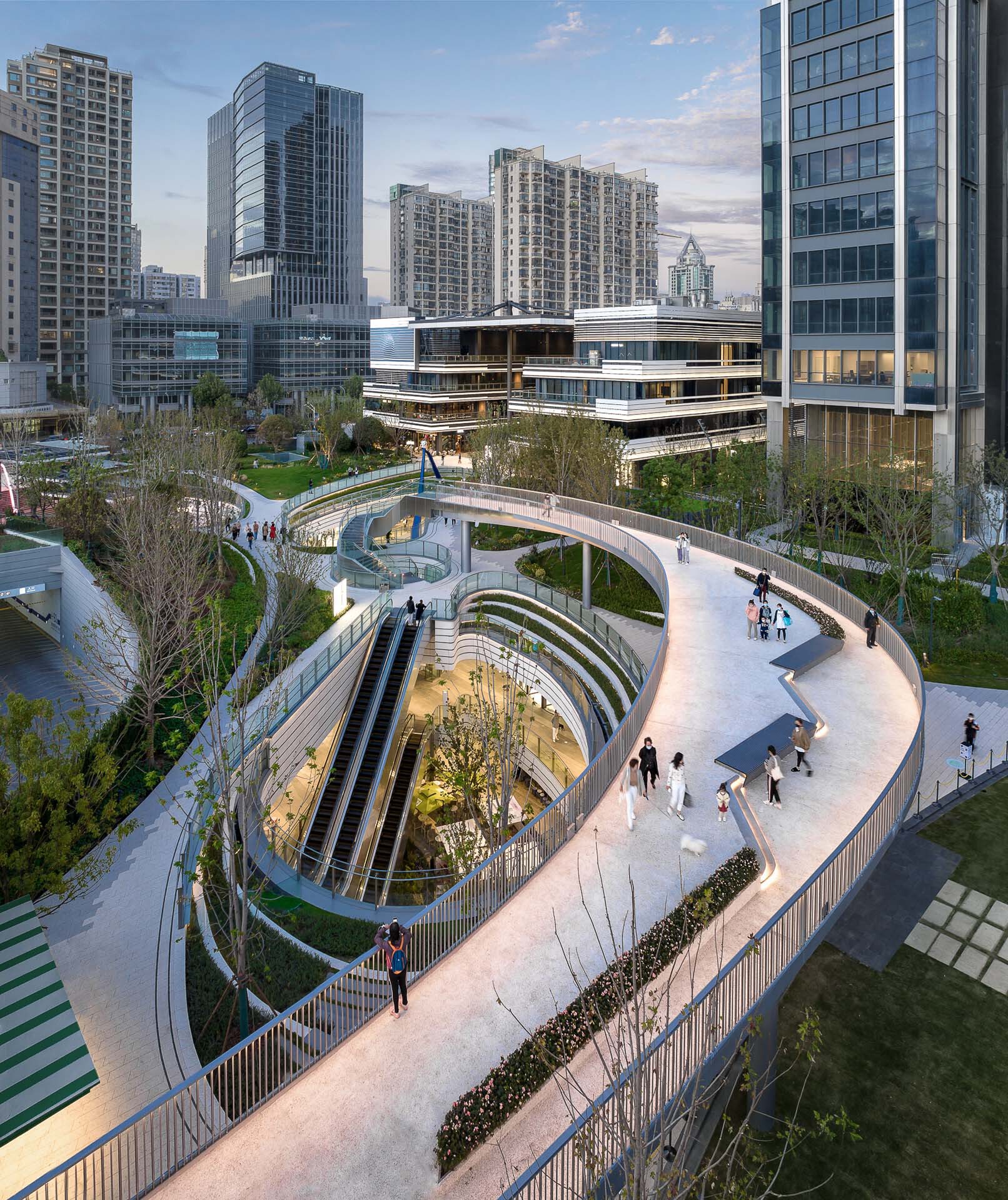 Spiraling pedestrian ramps descending around a sunken courtyard with trees at dusk