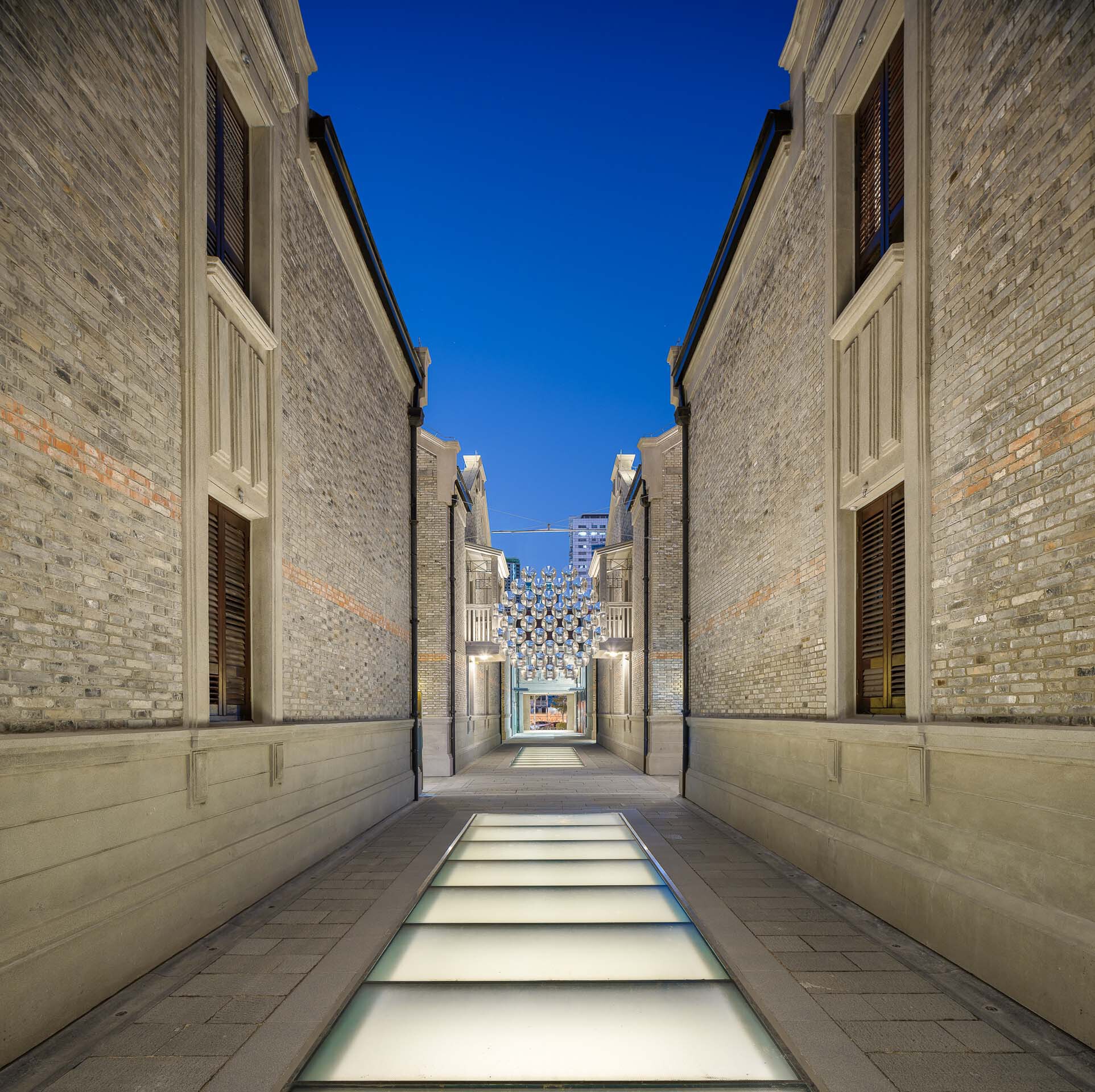Narrow lane with illuminated glass floor panels between grey brick walls at dusk
