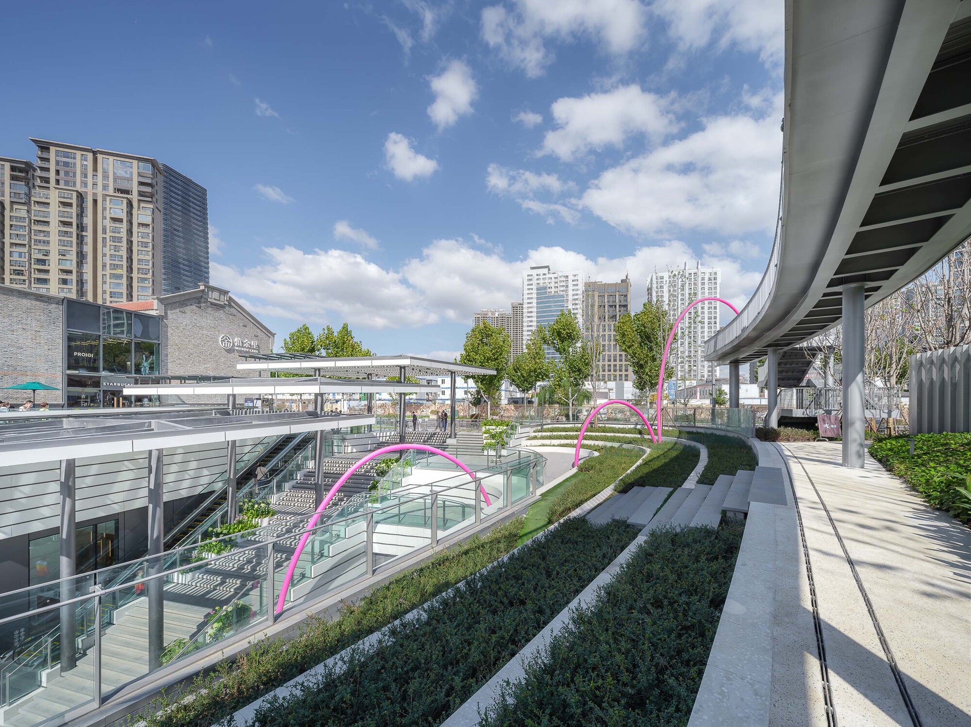 Elevated walkway with planted terraces and pink tubular sculpture beneath a highway overpass