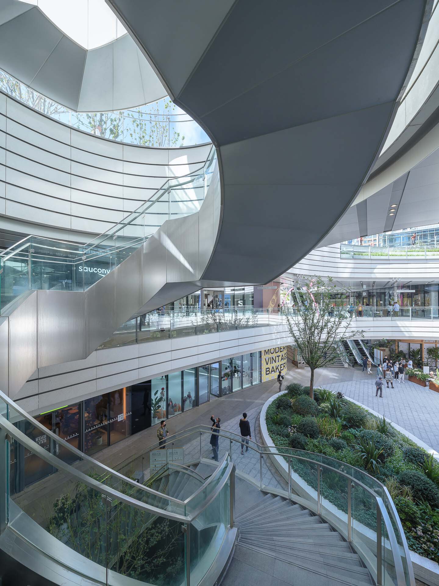 Multi-level atrium with curved white balconies, glass railings, planted terraces and shoppers on escalators below