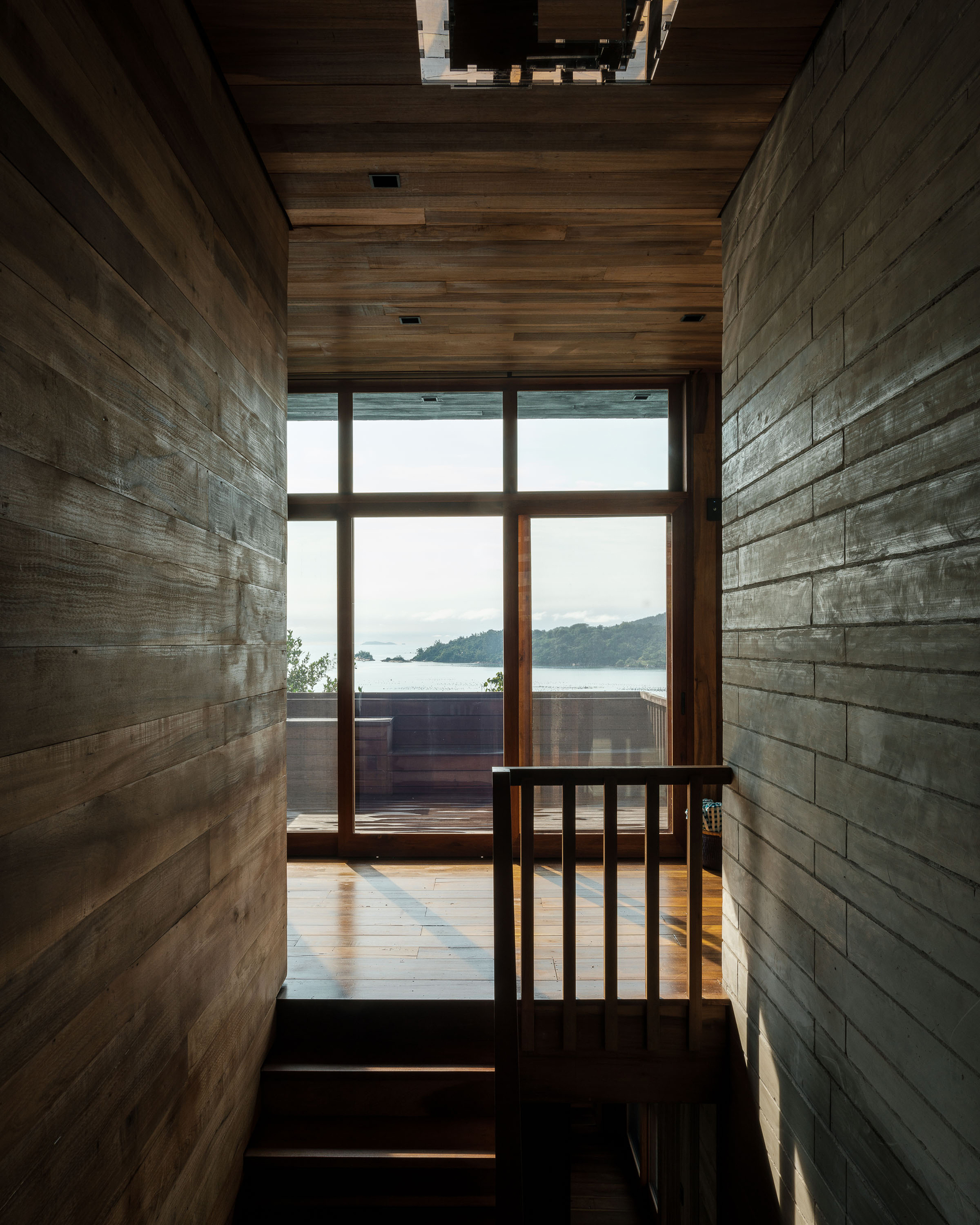 Narrow timber-clad corridor with board-formed walls framing a glazed door overlooking a coastal bay