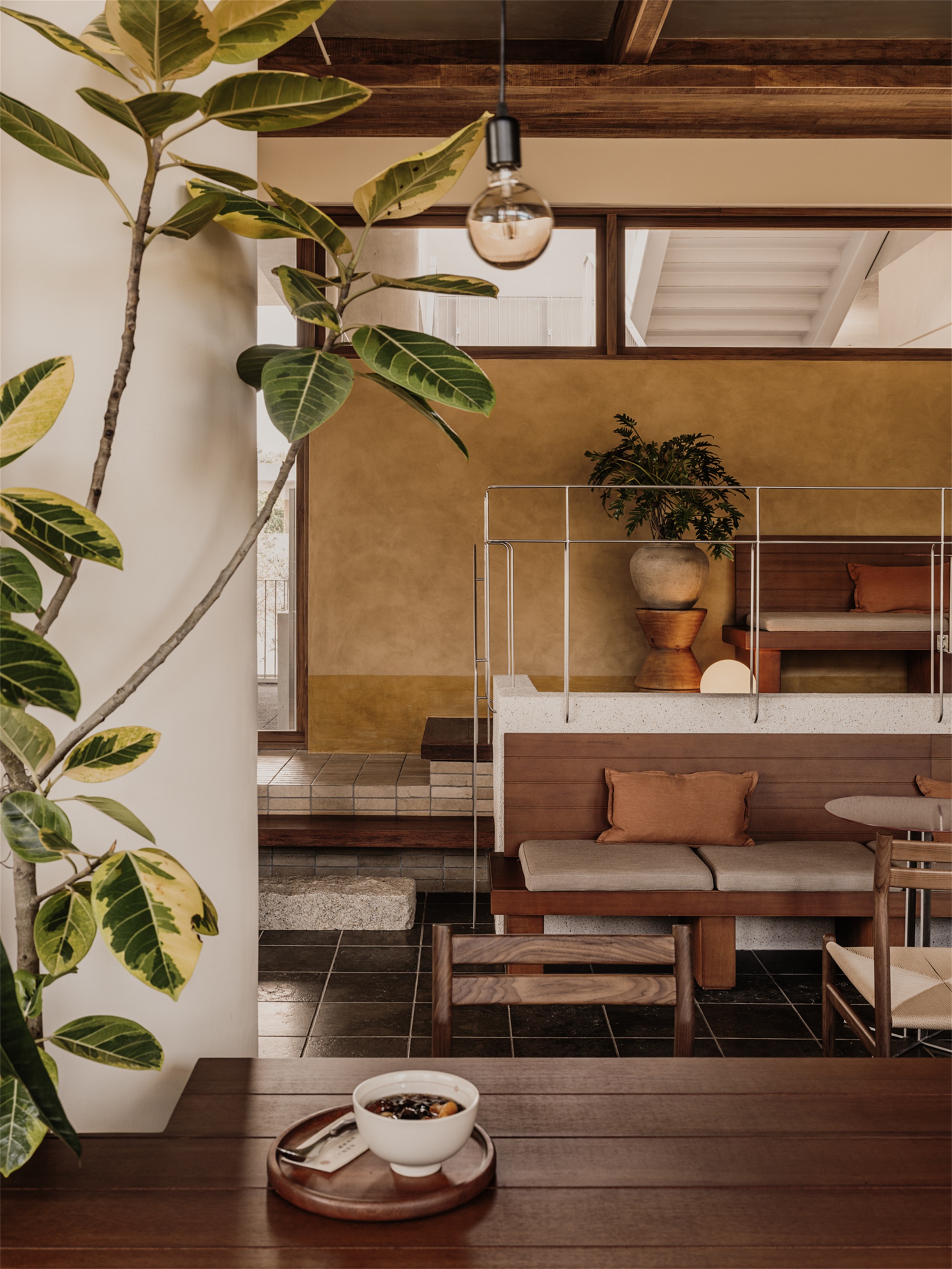 Interior view through timber-framed opening showing banquette seating, exposed ceiling beams, and potted rubber plant in foreground