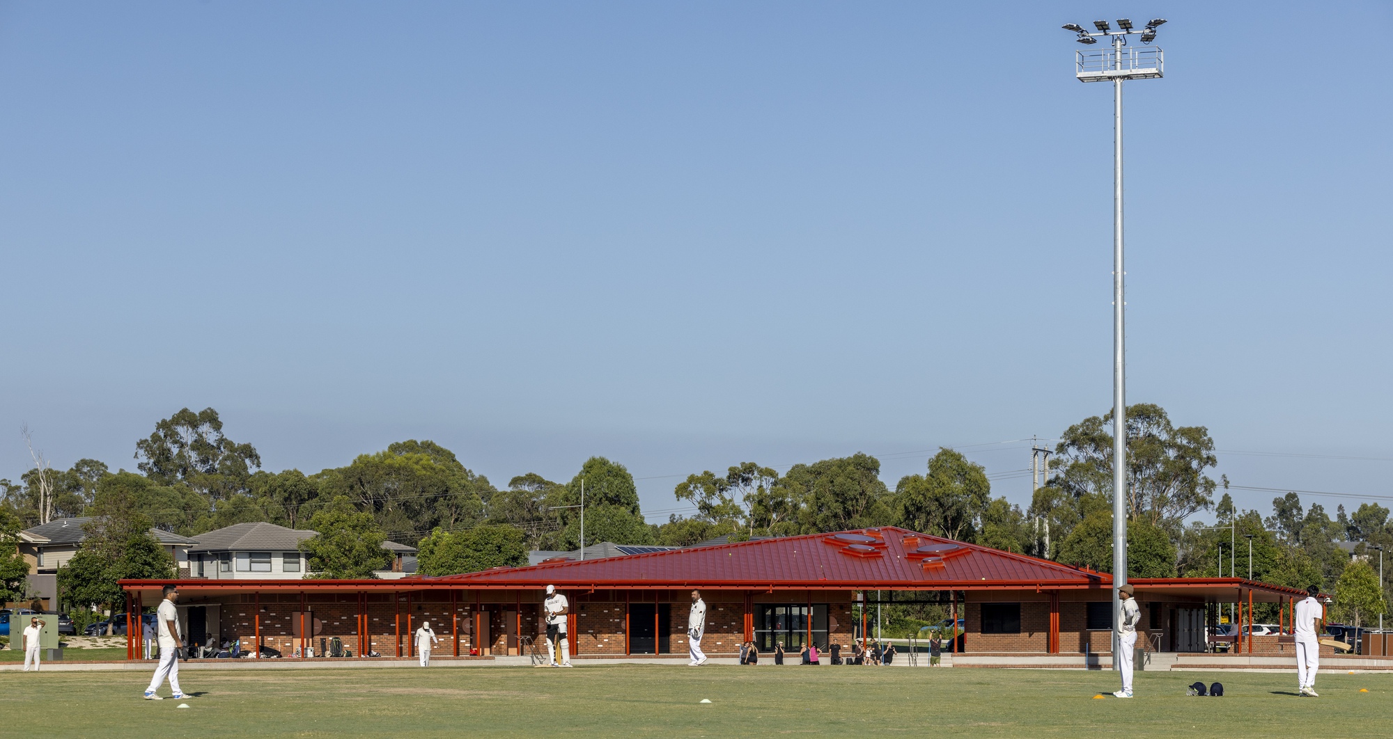 Long brick pavilion with red metal roof and deep eaves beside a lawn bowling green under clear sky