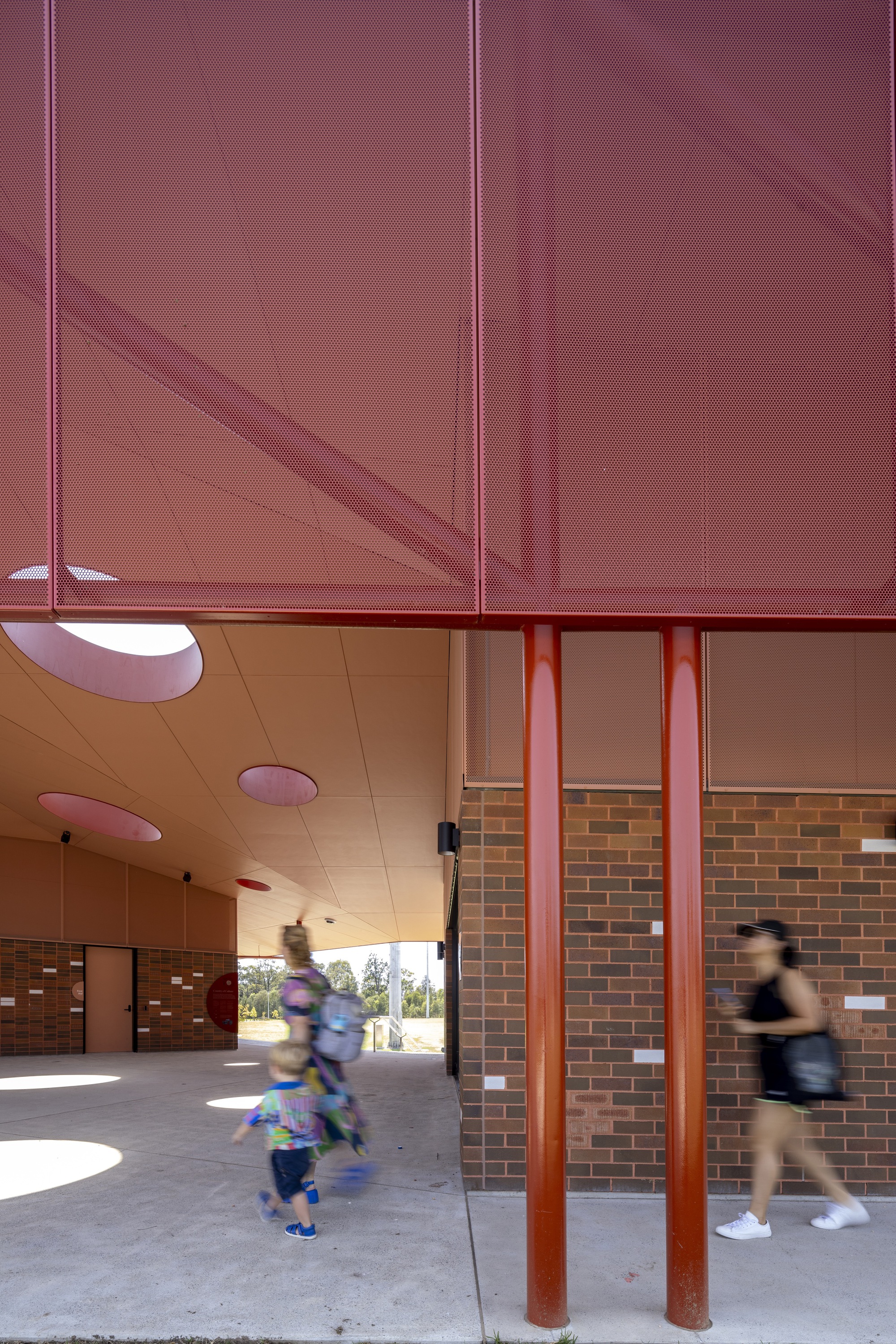 Covered breezeway with perforated red metal screen above and blurred figures walking through circular ceiling openings
