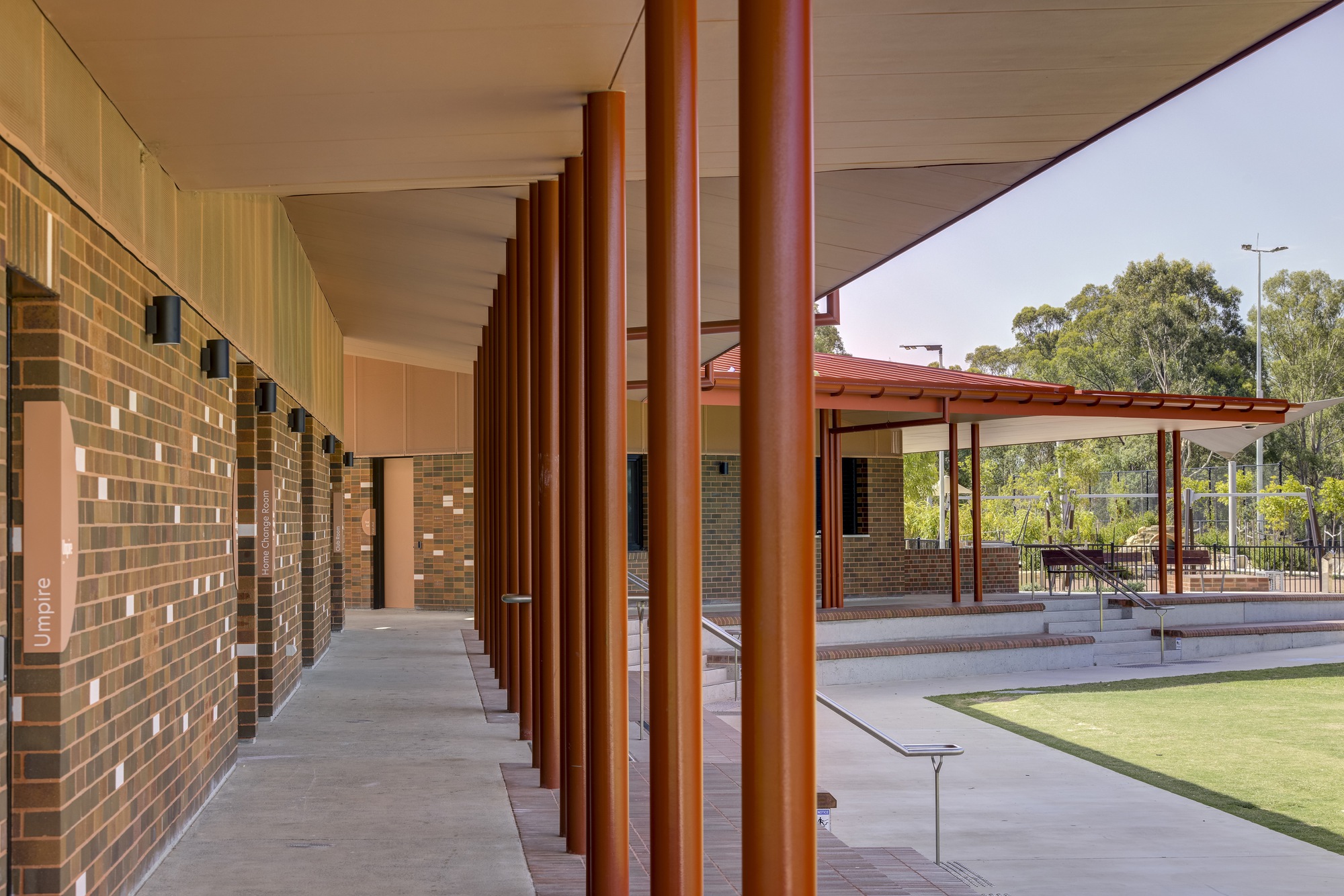 Covered walkway with reddish brown cylindrical columns along a multicolored brick wall under afternoon light